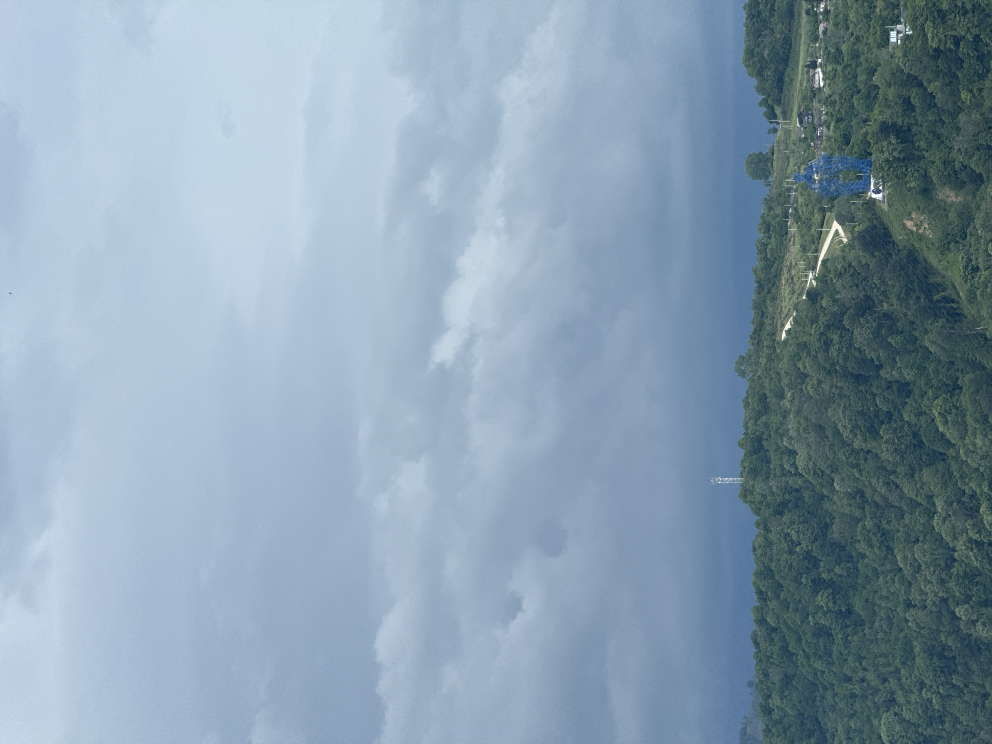 The image depicts a view in Sochi, Russia, with the sky taking up the majority of the frame. The sky is cloudy and gray, with varying shades of blue and white. On the right side of the image, there is a lush, green hillside covered with trees. Structures are visible on the ridge of the hillside. There is also a tall transmission tower visible.