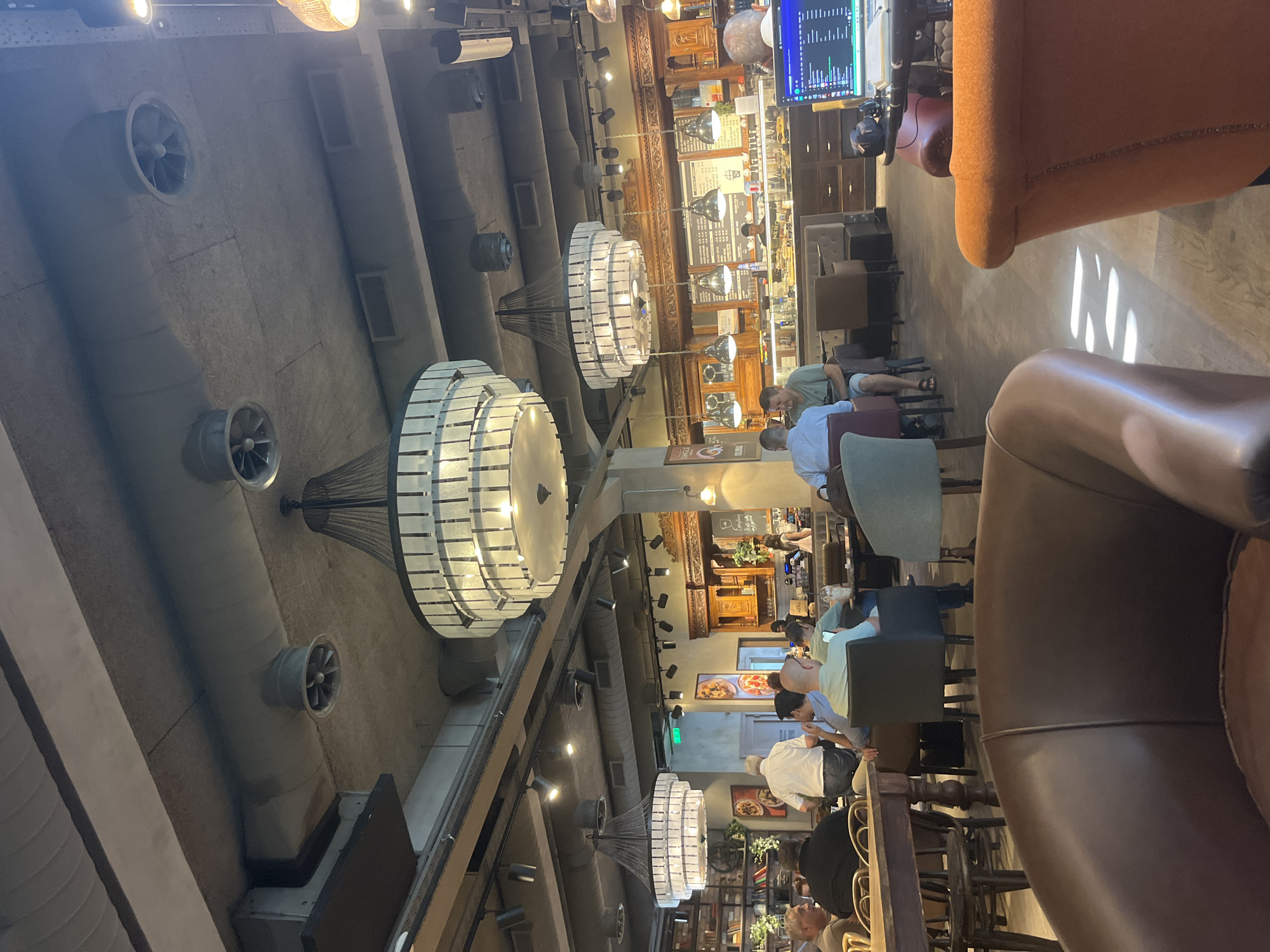 The image depicts the interior of a coffee shop, likely located in Agios Athanasios Municipality, Cyprus, based on the context provided. The setting is a bustling cafe with exposed concrete ceilings and industrial-style ductwork. Multiple large, ornate chandeliers hang from the ceiling, providing warm lighting.

Several patrons are seated at tables and in comfortable armchairs, engaged in various activities. In the foreground, two plush armchairs, one brown leather and one bright orange, are visible, suggesting seating areas. Further back, a group of people are seated at tables, some conversing while others look at their phones or laptops. One individual in a light blue shirt is looking at their phone. Another person in a green shirt is seated next to them, and a man in a white shirt is standing and looking towards the counter area.

The counter area features a display of food items and beverages. A barista can be seen working behind the counter. Menus are displayed on the wall behind the counter, indicating the services offered. The overall atmosphere appears relaxed and social, typical of a coffee shop environment. The flooring is made of light-colored wood planks. Sunlight streams in from unseen windows, casting geometric patterns on the floor. Bookshelves filled with books and plants are visible along one wall, adding to the decor.