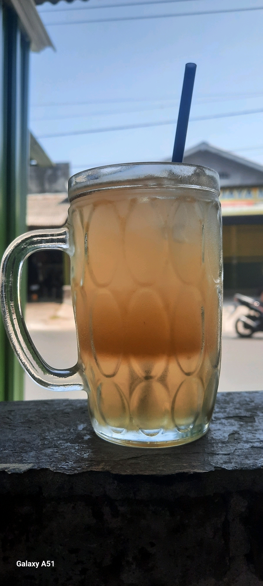 A tall, clear glass mug filled with iced tea and a blue straw is positioned on a textured concrete surface. The mug has a prominent handle and a patterned design. In the background, a blurry street scene is visible, with a motorcycle parked on the right side. The sky above is clear and blue, with a hint of cloud. The foreground shows the edge of the concrete surface. The image was taken in Selopuro, Indonesia, as indicated by the "Galaxy A51" watermark, suggesting it was captured with a Samsung Galaxy A51 phone. The lighting suggests it is daytime.