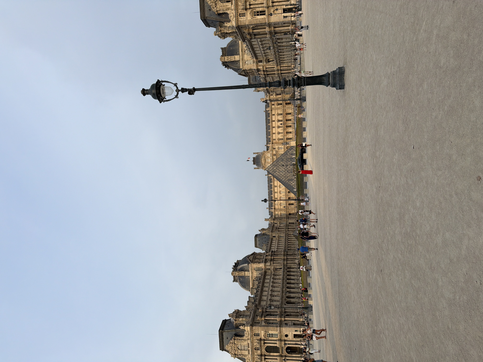 The image depicts the exterior of the Louvre Museum in Paris, France, with the iconic glass pyramid visible in the background. The foreground is a wide, open plaza with a gravel surface. Several people are scattered across the plaza, walking and standing. A prominent black lamppost stands in the right-middle of the frame, its intricate design silhouetted against a pale sky. The Louvre building itself, a grand architectural masterpiece with ornate facades and dark, mansard roofs, stretches across the mid-ground. A French flag flies atop one of the building's structures. The scene appears to be captured during daylight, possibly late afternoon or early evening, given the soft lighting and the clear sky.