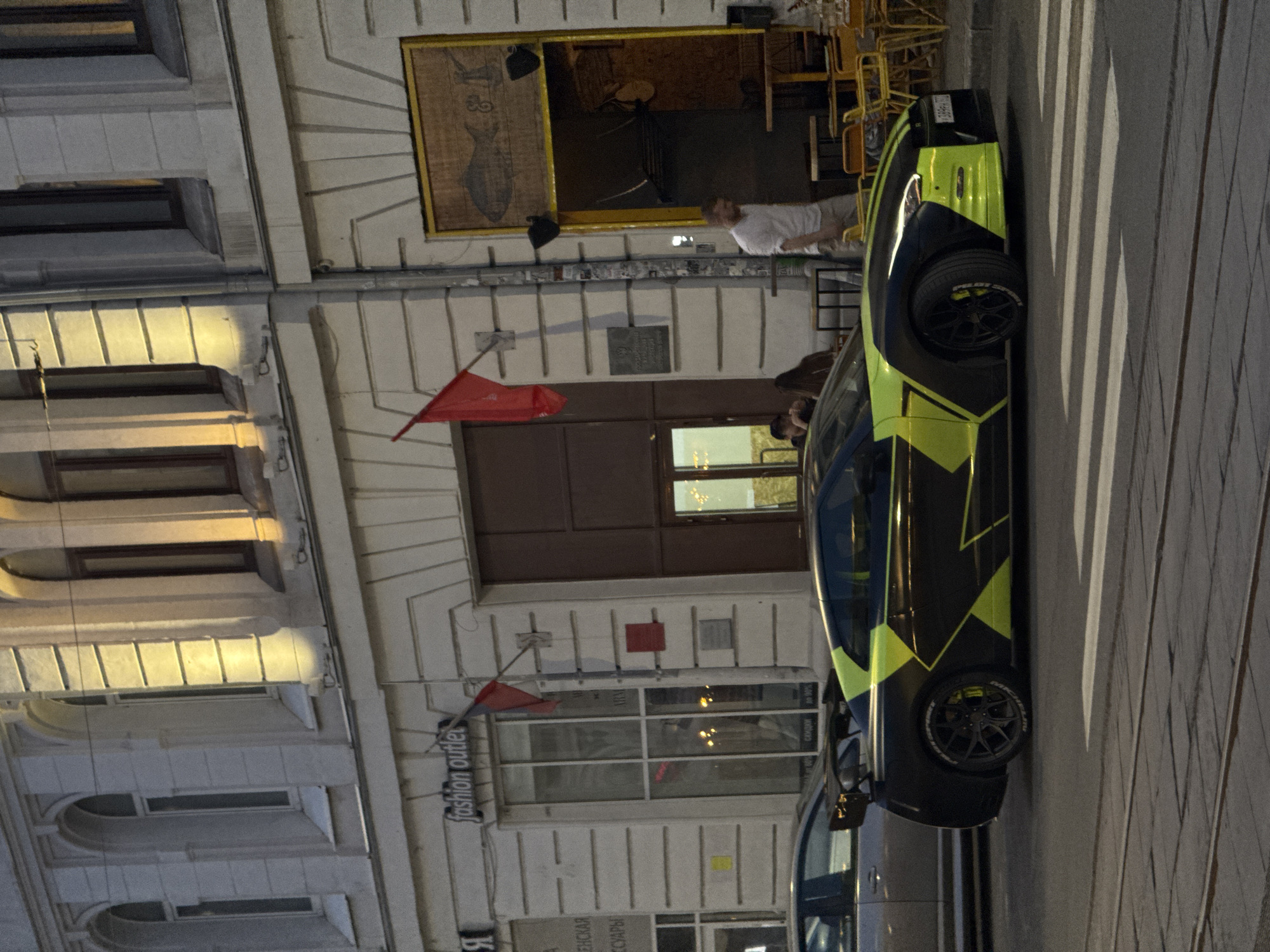 The image captures a street scene in Nizhny Novgorod, Russia. A modern car with a striking black and lime green color scheme is parked on the right side of the frame, taking up a significant portion of the visual space. The street pavement appears to be made of cobblestones.
Buildings line the street, displaying a mixture of architectural styles. A red flag is visible on one of the buildings, and signage indicates a "fashion outlet". A store or establishment with a wooden-framed opening and seating can be seen in the background, with a man standing near the entrance.
The lighting suggests it is either late evening or early morning. There are a few people present in the scene, including the man by the establishment and others partially visible near the car. There are no visible actions or interactions beyond the presence of people and parked vehicles.