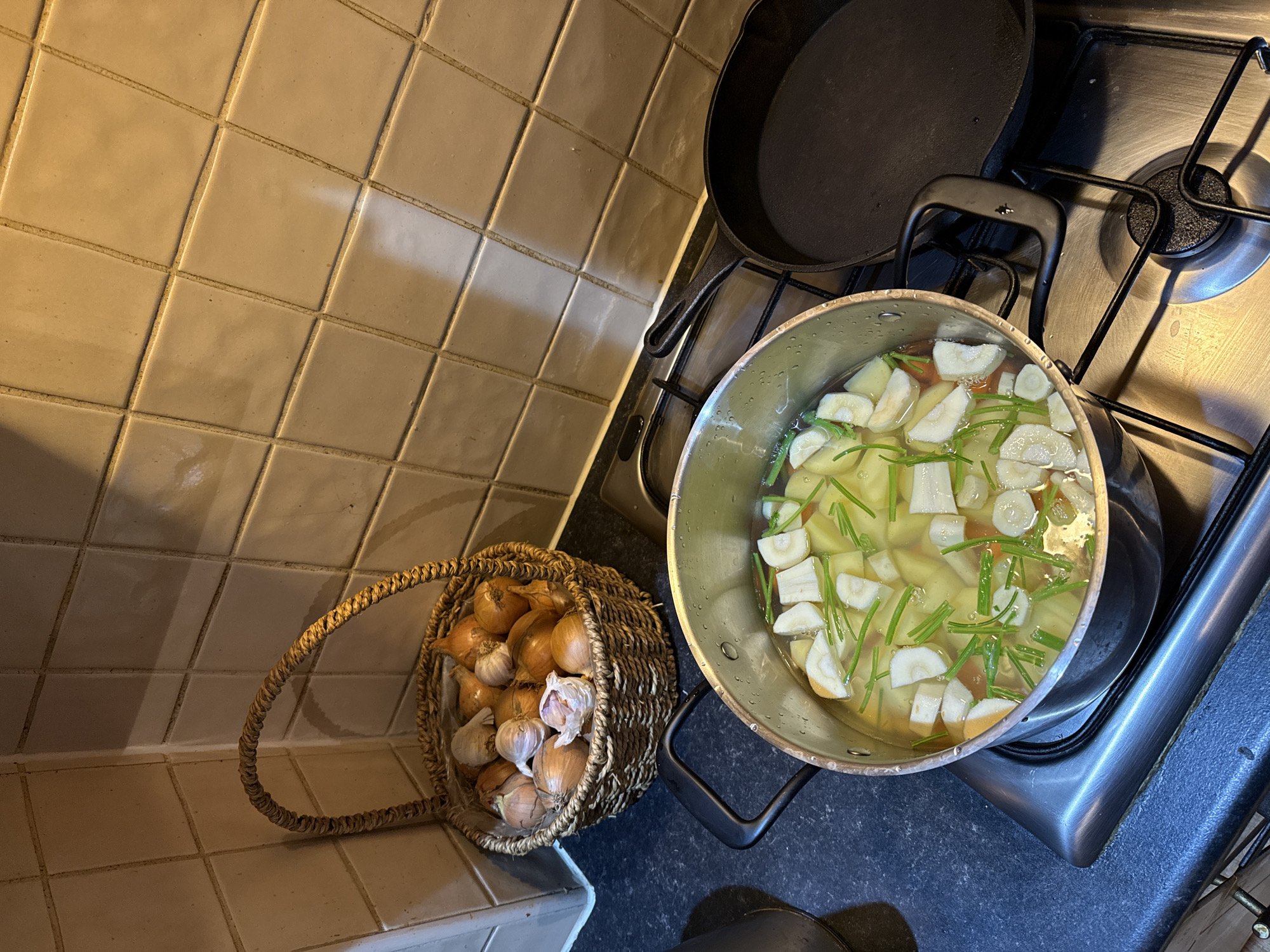 The image depicts a kitchen scene in Great Yarmouth, United Kingdom. A woven basket filled with onions and garlic sits on a tiled countertop. Next to it, a stainless steel pot rests on a gas stovetop, filled with water, potatoes, parsnips, carrots, and green herbs (possibly leek greens or chives). A black cast iron skillet is positioned near the pot on the stovetop. The countertop and stovetop appear to be made of a dark material. The wall behind the countertop is tiled with light beige squares. No people are visible in the image. The scene suggests a meal is being prepared.