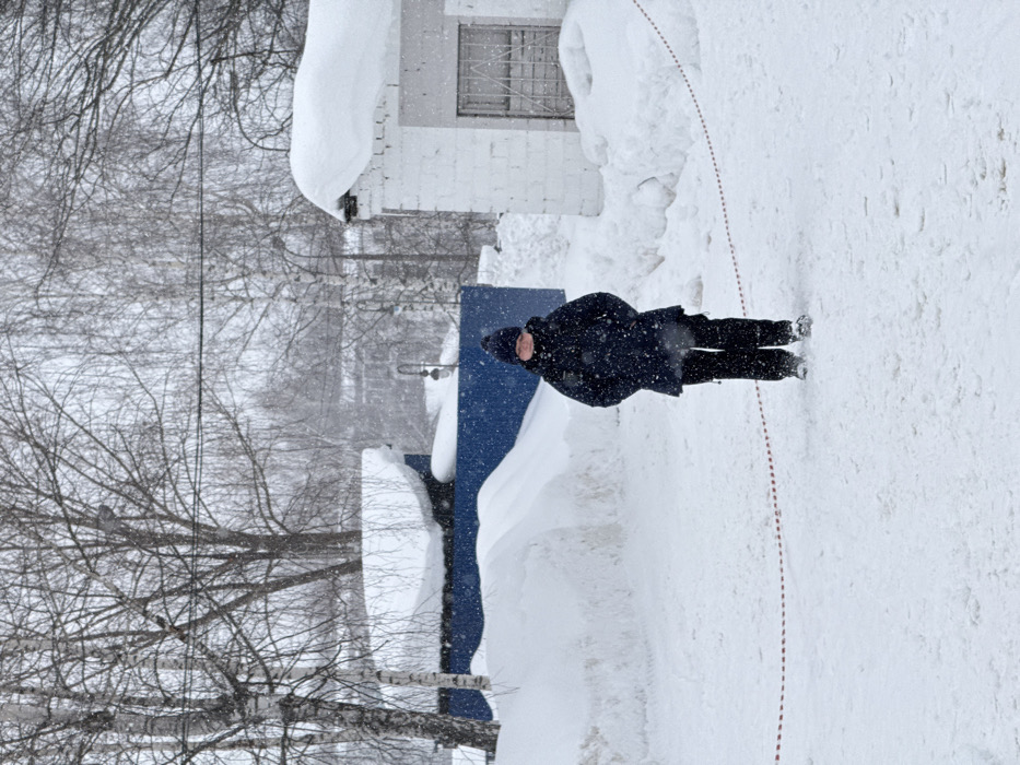 The image depicts a winter scene in Chaikovsky, Russia, characterized by heavy snowfall. A single individual, appearing to be a child or young adolescent, stands centrally in the frame, facing forward. This person is dressed in a dark blue/black winter coat, dark pants, and a dark knitted hat. Snowflakes are visible on their clothing and in the air. The ground is covered in a thick layer of undisturbed white snow, with some footprints visible. A segmented red line or tape lies horizontally on the snow in the lower portion of the image, curving slightly.
To the left of the individual, partially obscured by snowdrifts, is a dark blue structure, possibly a container or small building, with its roof heavily laden with snow. To the right, a white brick building with a dark roof, also heavily covered in snow, features a gridded opening or window. Large snowdrifts are accumulated against these structures and along the perimeter of the open area. Bare deciduous trees, including several birch trees with visible bark, are prominent in the midground and background, their branches outlined against an overcast sky. Numerous snowflakes are visibly falling throughout the scene.