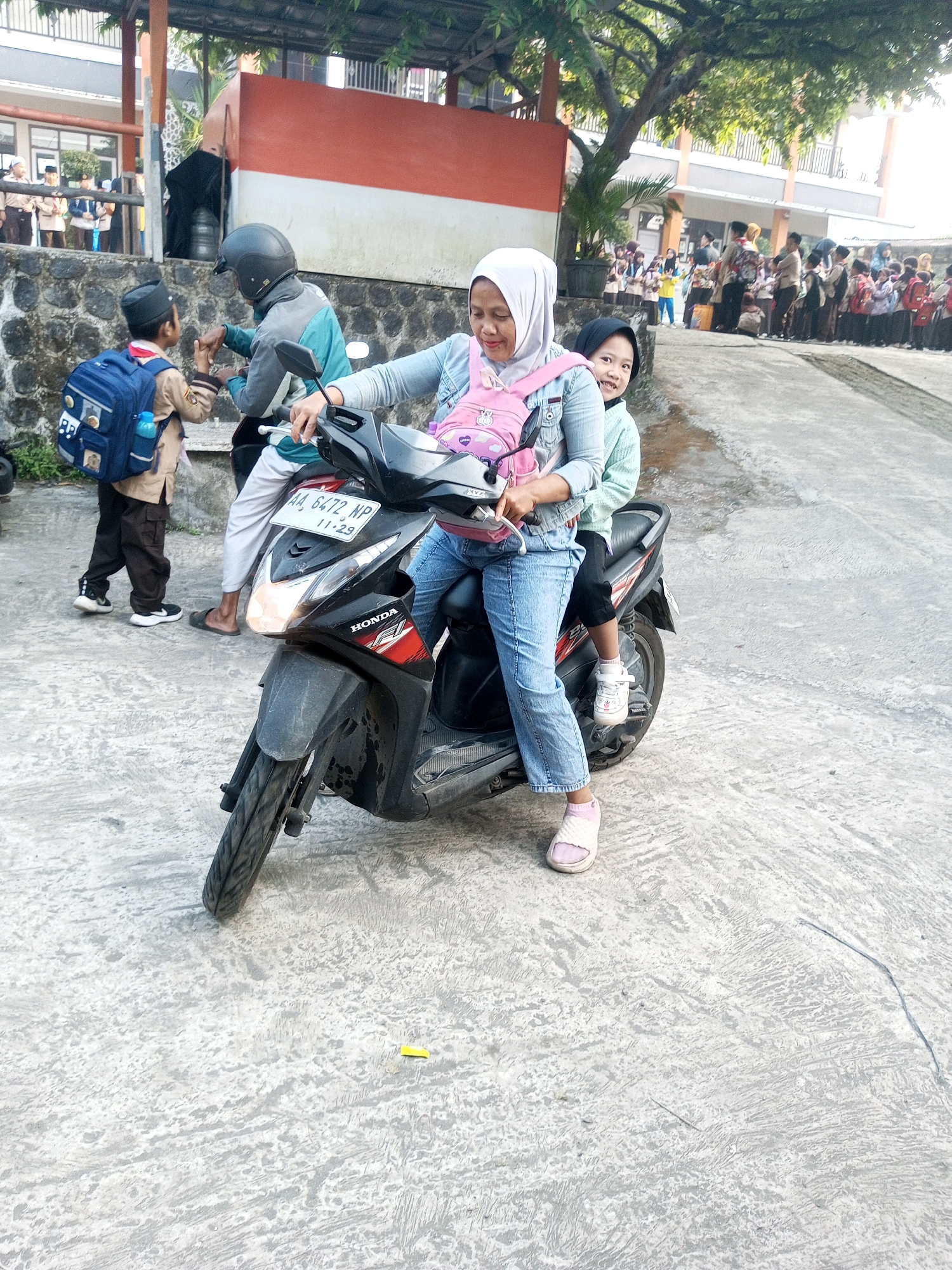 In Krasak, Indonesia, a woman wearing a light blue denim jacket, blue jeans, and a white hijab rides a black Honda scooter. She is holding the handlebars, and a young child, wearing a light green sweater and black pants, is sitting behind her. The child is smiling and looking towards the camera. The scooter has a license plate that reads "AA 6472 NP 11-29". To the left of the scooter, a young boy in a scout uniform and a blue backpack is talking to a person wearing a helmet and a teal jacket. In the background, a group of students in uniform are lined up, and a tree with green foliage is visible. The scene appears to be in front of a building with red and white accents. The ground is paved with concrete.