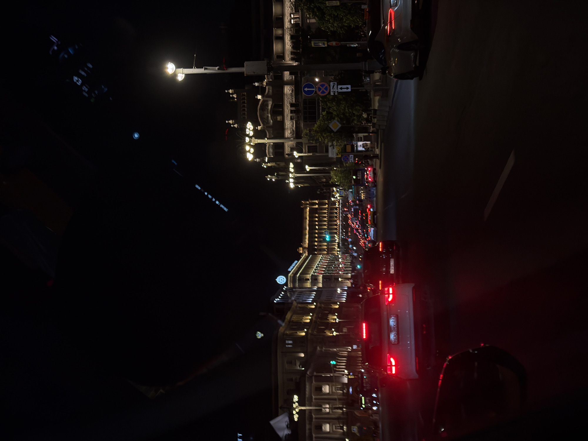 The image shows a night view of a street in Moscow, Russia. Traffic is heavy with numerous cars visible, their taillights illuminated. Streetlights and building lights add to the overall brightness of the scene, particularly highlighting ornate architectural details. Road signs and traffic signals are also visible.