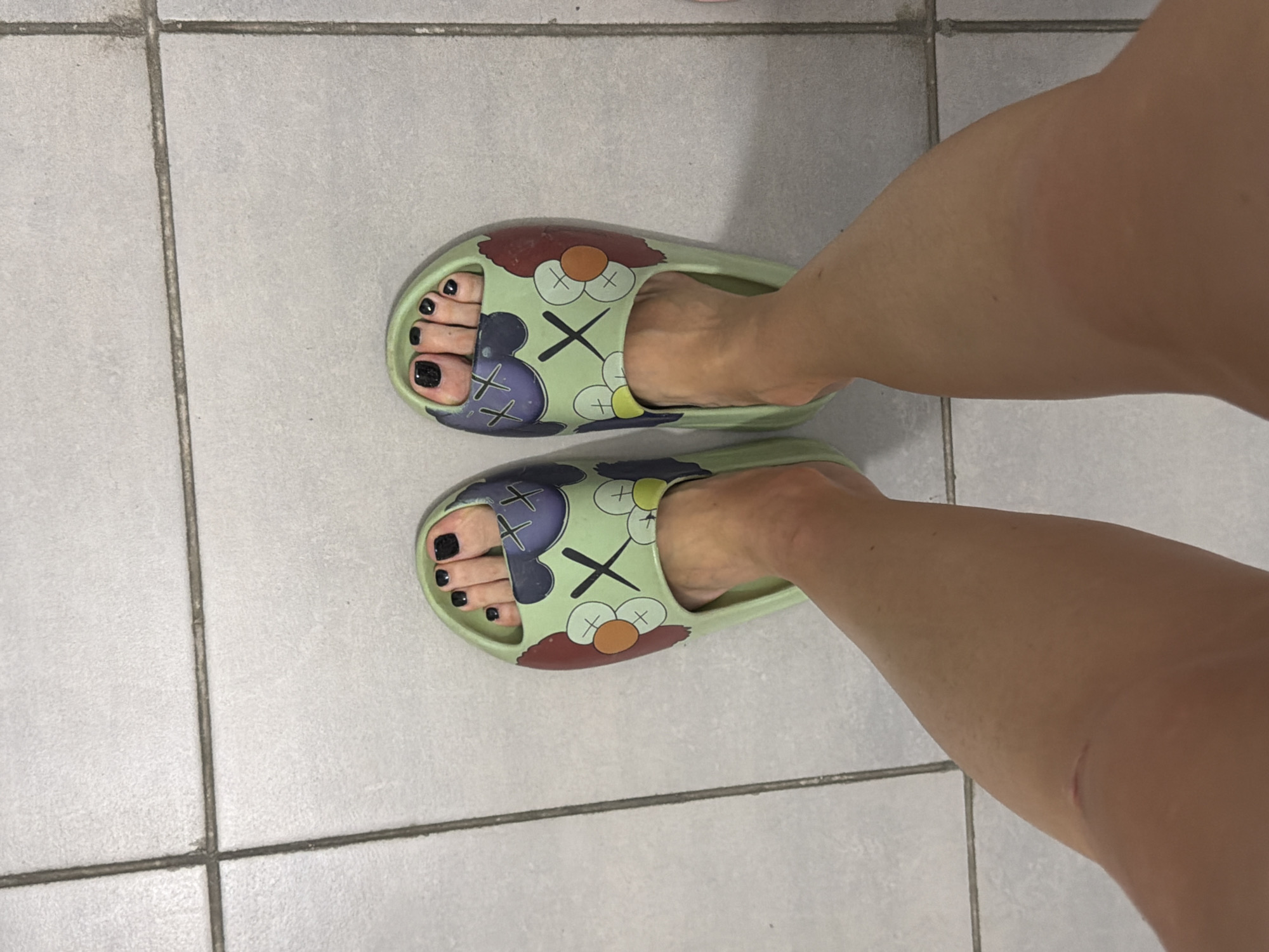 The image shows a pair of feet wearing light green slide sandals with a cartoonish design featuring "X" eyes and circular shapes. The toenails are painted black. The feet are standing on a tiled floor, with grout lines visible. The floor tiles are square and light gray. The lighting appears to be artificial. The image is a close-up, waist-down view of the subject. The location in Novoalexandrovka, Russia is not discernible from the visual information provided.