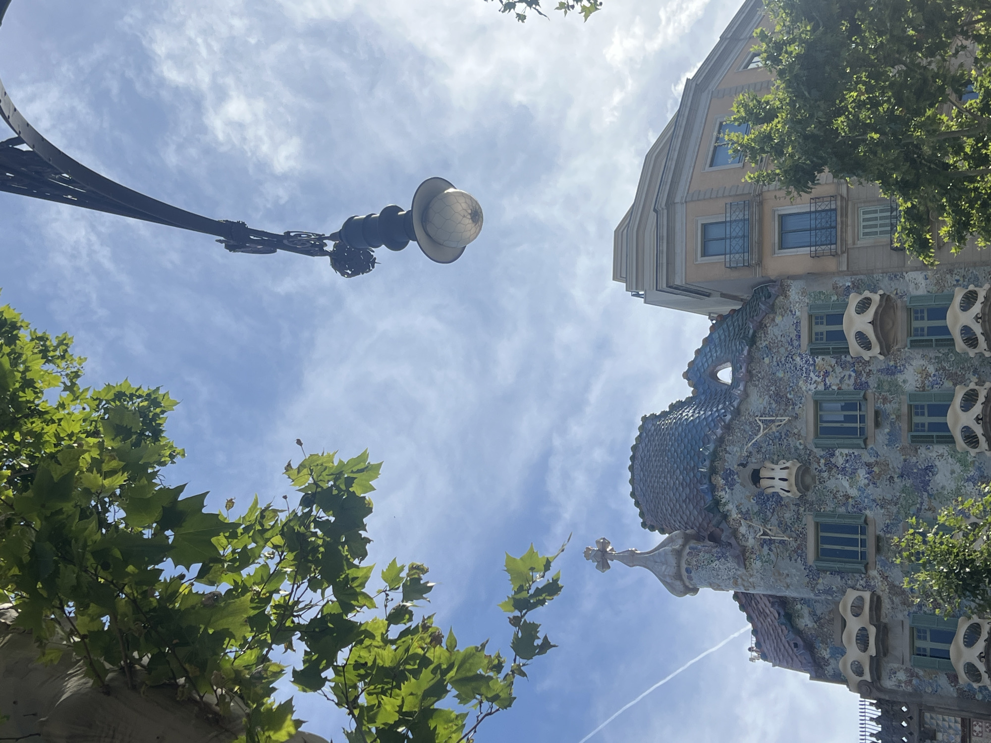 This is a low-angle, full-frame shot of Casa Batlló in Barcelona, Spain, on a bright, cloudy day. The building's facade is covered in colorful mosaics and sculpted elements, with a distinctive dragon-scale roof. A tall, ornate lamppost with a glass globe hangs from the top right corner of the frame, silhouetted against the sky. Lush green trees with broad leaves frame the top left and right sides of the image. The sky is blue with scattered white clouds, and a faint contrail is visible.