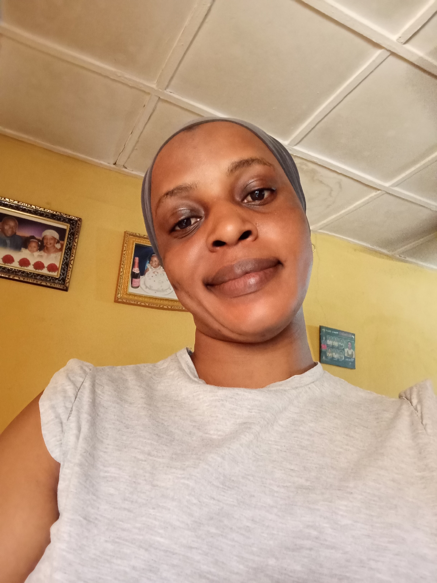 A medium, eye-level shot shows a woman wearing a gray head covering and a gray short-sleeved shirt. The background is a yellow wall with three framed pictures. The ceiling is white with square tiles. The location is likely a residence, possibly in Narayi, Nigeria.