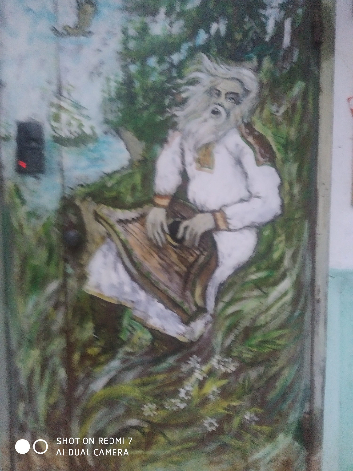 The image shows a painting on a wall in the city of Chaikovsky, Russia. The painting depicts a man with long white hair and a beard, wearing a white shirt with embroidered sleeves and playing a harp. The man is surrounded by green foliage and white flowers. In the background, there is a body of water, trees, and a bird in flight. To the left of the painting, there is a black intercom box with a red light. The bottom of the image contains the text "SHOT ON REDMI 7 AI DUAL CAMERA".