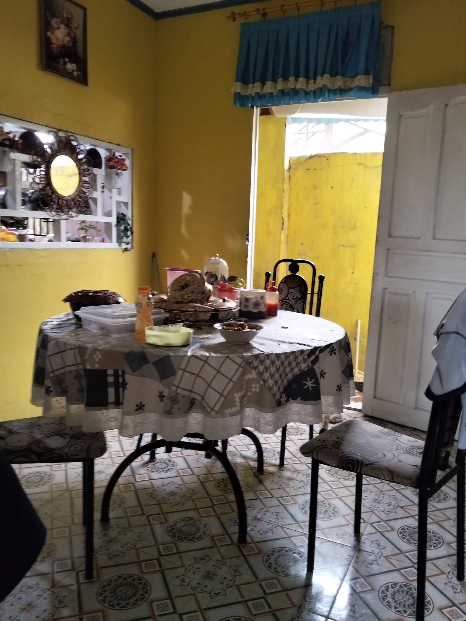 The image depicts a dining table set for a meal in a room with yellow walls and tiled flooring. The table is covered with a patterned tablecloth and laden with dishes, including bowls of food, a bottle of sauce, and a container. Two chairs, one with a patterned seat and the other a black metal chair, are placed around the table. A doorway leads to an outdoor area with a yellow wall and a white door. On the wall, a framed floral picture hangs above a mirror with a decorative frame. A blue curtain with ruffles is visible near the ceiling. The scene appears to be an indoor dining area in a home located in Arang Limbung, Indonesia.