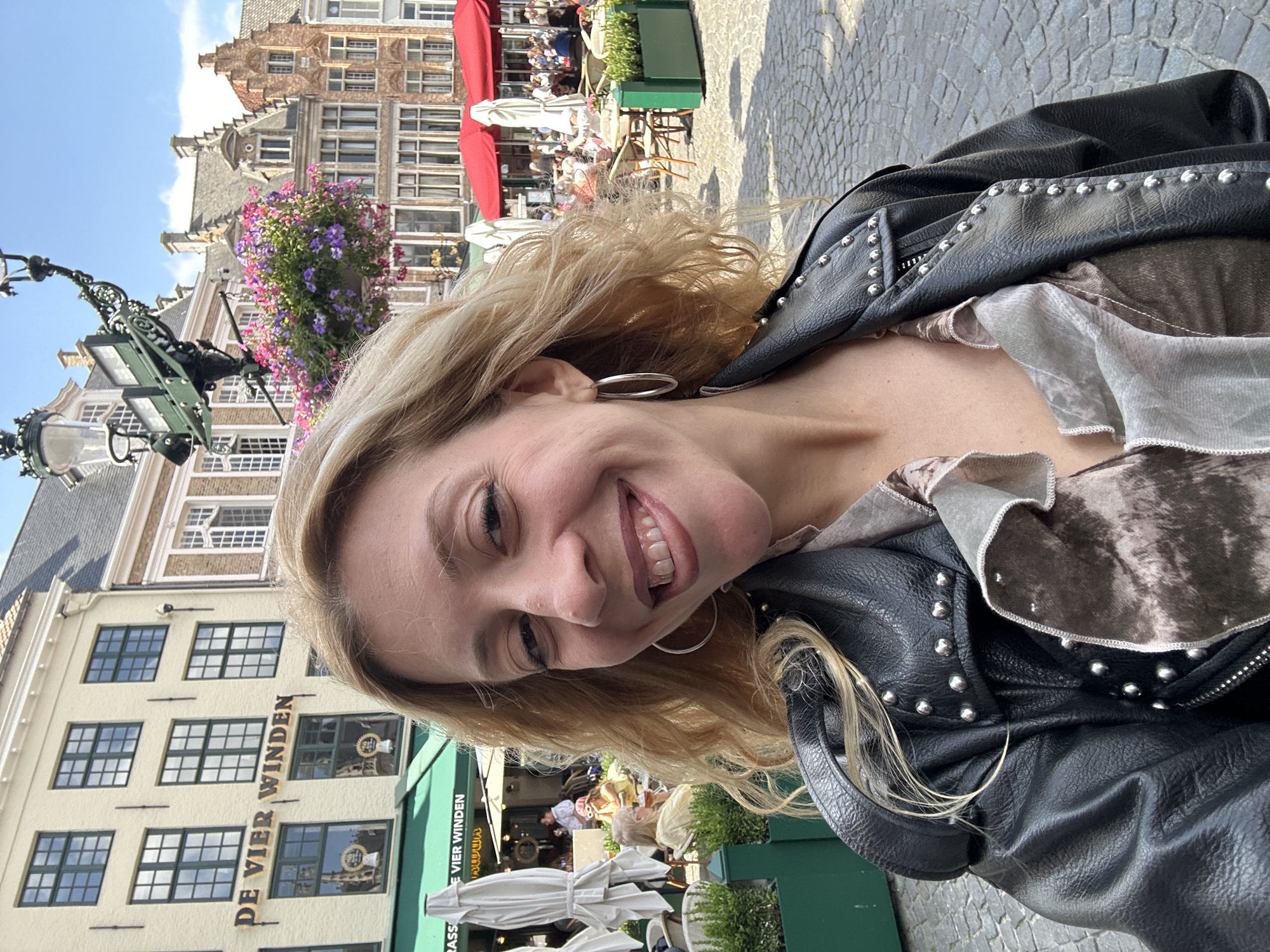 This is a medium-shot, selfie-style photograph of a fair-skinned, blonde woman with light-colored eyes and a wide smile. She is wearing a black leather jacket adorned with silver studs over a patterned grey and brown silk blouse. Her hair is wavy and falls around her shoulders. She is wearing large silver hoop earrings.
The background is a cobblestone plaza in Bruges, Belgium, bathed in sunlight under a partly cloudy sky. Behind her, the buildings are typical of Bruges architecture, with gabled facades and cream-colored walls. A sign on one building reads "DE VIER WINDEN". Red and white umbrellas are set up outside an establishment, with tables and chairs visible, and several patrons seated. A hanging basket overflowing with pink and purple flowers is suspended from a lamppost. Green planters are placed around the plaza. The overall scene depicts a vibrant, outdoor urban setting.