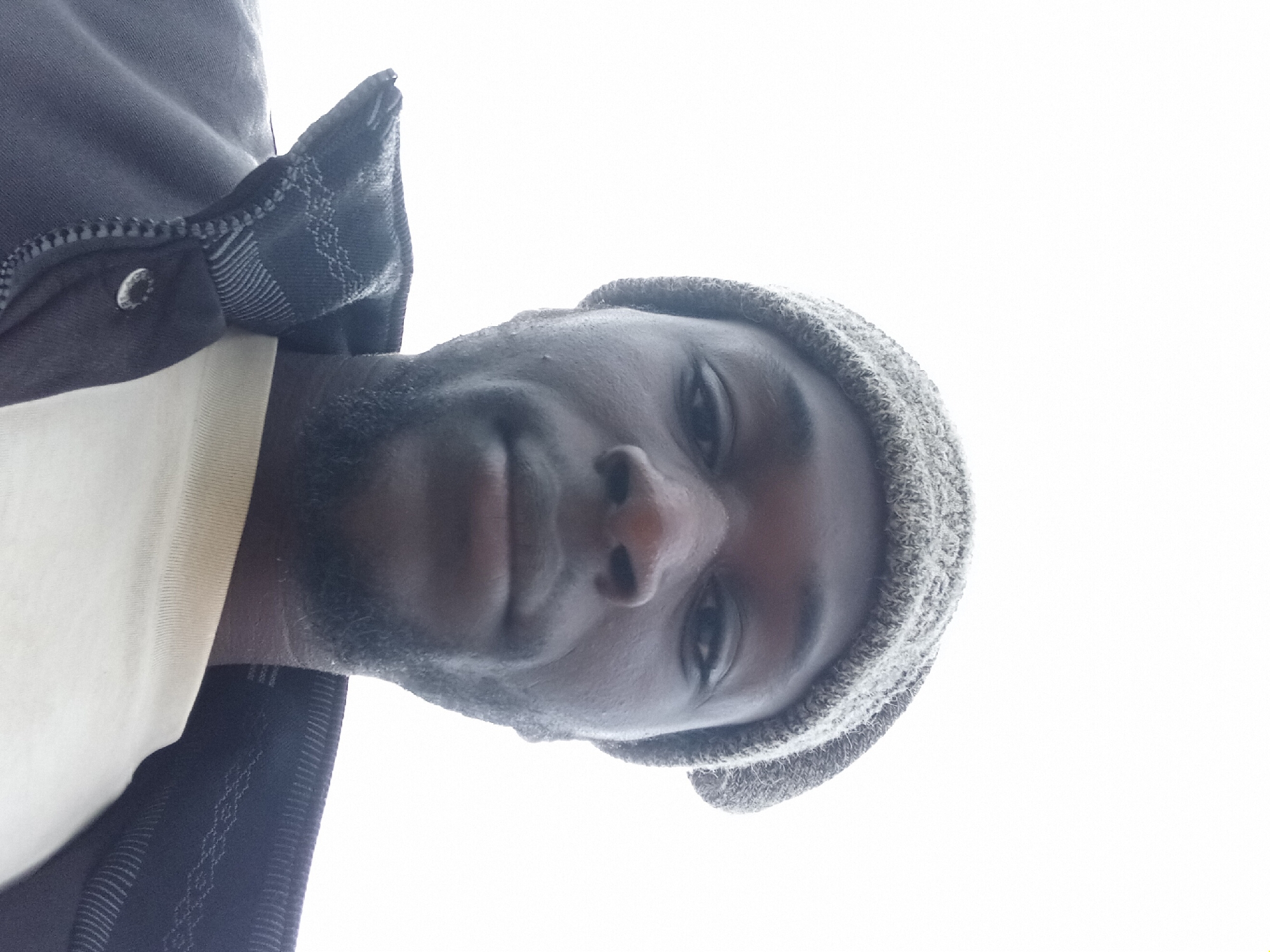A low-angle, close-up, full-frame selfie of a Black man wearing a grey knit beanie and a dark jacket over a light-colored t-shirt is taken against a bright, featureless sky, suggesting an outdoor environment in Bukuru, Nigeria. The man's head is tilted slightly upwards, and he is looking directly at the camera. His skin appears dark, and he has short, neatly trimmed facial hair. The lighting is harsh and comes from above, creating a blown-out effect in the background and strong shadows on his face. The focus is sharp on his face and upper body, highlighting the texture of his skin, the knit of his hat, and the fabric of his clothing. The image is candid and informal, characteristic of a selfie.
