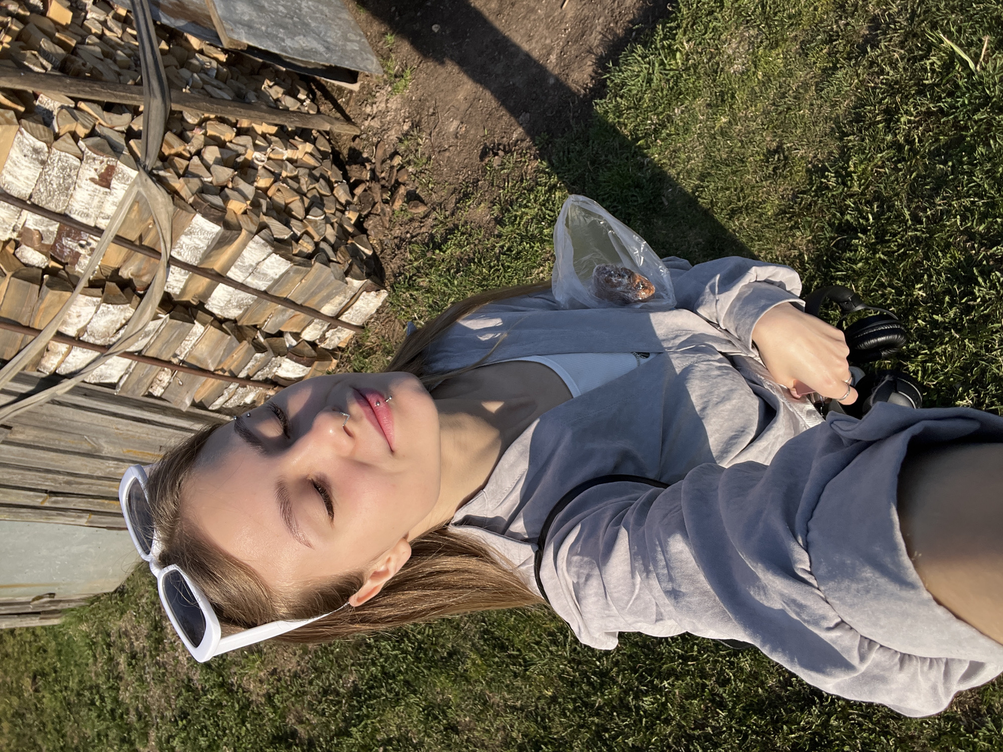 A young woman with fair skin and light brown hair is lying on a patch of green grass, with her head near a stack of firewood. She is wearing sunglasses, a white tank top, and a gray sweatshirt. In her right hand, she holds black headphones. A plastic bag containing a pastry or bread sits near her left shoulder. The environment appears to be a rural or semi-rural outdoor setting, likely in Фоки, Russia, characterized by a mix of natural elements (grass) and man-made structures (woodpile, possible building elements in the background). The scene is bathed in sunlight, suggesting a day-lit environment. The woman's eyes are closed, hinting at relaxation or rest.