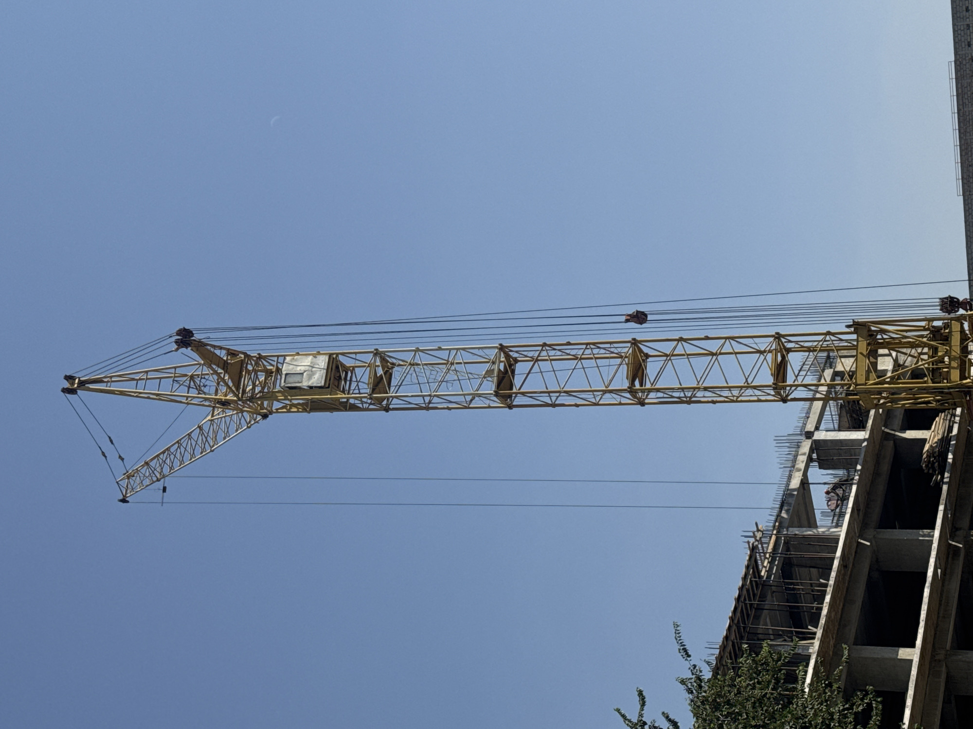 A tall yellow construction crane stands against a clear blue sky, positioned to the right of a partially constructed concrete building. The building has multiple floors with exposed rebar and scaffolding. Some trees with green foliage are visible on the left side of the frame, partially obscuring the building. The crane's cab is visible near the top of its vertical tower. The scene appears to be a construction site in the city of Bishkek, Kyrgyzstan.