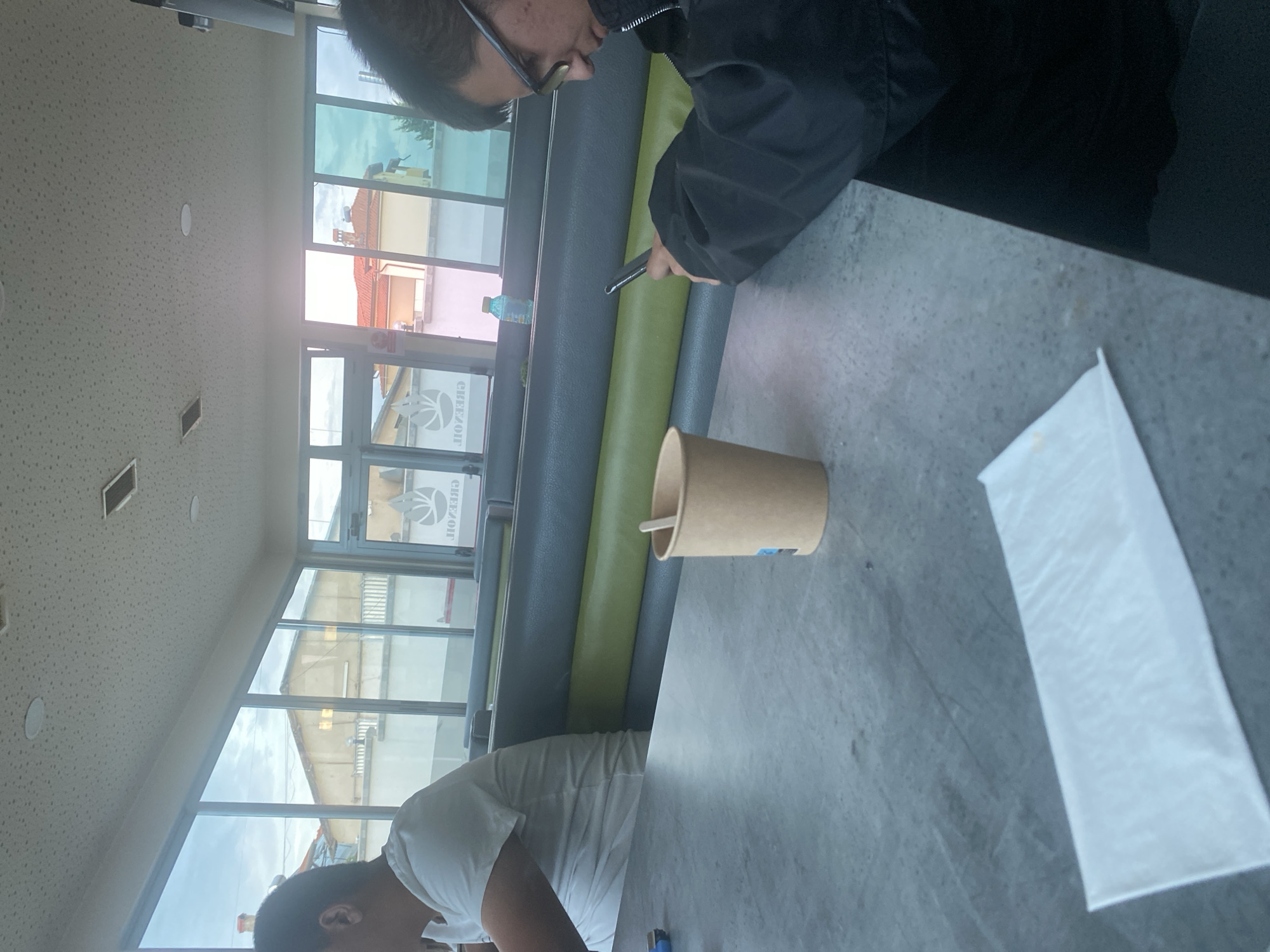 In Ablanitsa, Bulgaria, two individuals are seated at a table within a diner. The person on the left, facing away from the camera, wears a white t-shirt. The person on the right, in profile, wears glasses and a black jacket, engrossed in their smartphone. A brown paper cup with a wooden stirrer sits on the table, along with a white napkin and a blue lighter. The diner features large windows overlooking buildings and a cloudy sky, with green and gray booth seating lining the interior.