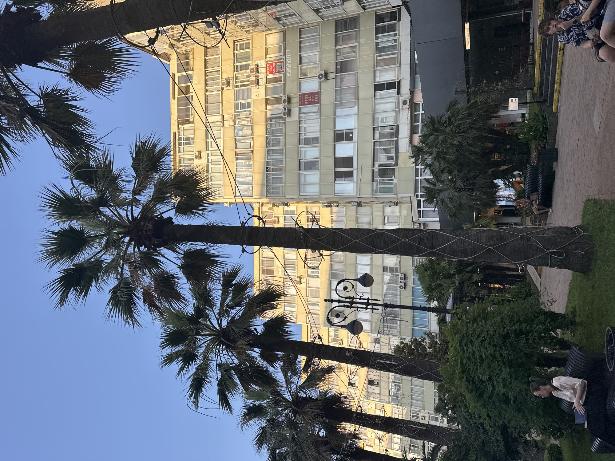 The image depicts a street scene in Sochi, Russia, characterized by tall palm trees framing a multi-story, light-yellow apartment building. The building features numerous windows, balconies, and air conditioning units, suggesting a residential or mixed-use urban environment. A vintage-style street lamp with two globes stands prominently in the foreground, partially obscured by the palm trees.
In the lower portion of the image, a paved walkway and grassy area are visible. Two individuals are seated on a dark bench in the grassy area on the left, with one person, a woman wearing a light-colored top and dark pants, holding a book. Another group of people is seated on benches in the background, near a dark awning with "EAZZY COFFEE" signage. The sky above is clear and blue, indicating daytime. The overall impression is one of a tropical urban setting, likely during warm weather.