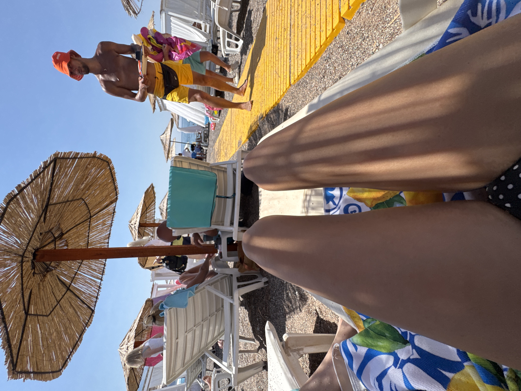 In a beach setting, two individuals are seated on lounge chairs, their legs prominently displayed in the foreground. The chairs are covered with colorful towels, one featuring a lemon pattern. Behind them, several more lounge chairs and straw umbrellas are visible, suggesting a resort or public beach area. In the background, a man wearing yellow shorts and an orange hat walks on a yellow boardwalk, carrying some items. The sky is clear and blue, indicating a sunny day. The overall environment suggests a vacation or leisure activity on a beach.