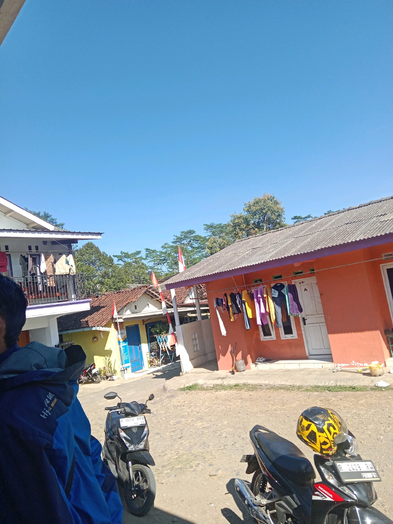 In Leksono, Indonesia, a sunny day is captured with a clear blue sky dominating the upper half of the image. In the foreground, two motorcycles are parked on a dirt and gravel surface. The motorcycle on the left is black with a license plate reading "R 3652 DAD". A person wearing a blue jacket is partially visible in the left foreground, their back to the camera. On the right, another black motorcycle has a yellow and black patterned helmet resting on its handlebars, and its license plate displays "R 2473 RH".
Behind the motorcycles, several houses are visible. The house on the right is painted an orange hue and has laundry hanging on a line in front of it, including shirts, pants, and other garments. Two white-framed windows are present below the clothesline, and a white door is to the right of the windows. The house on the left features a white facade with a balcony on the second floor, where more clothes are hanging. Red and white Indonesian flags are visible, attached to poles in front of the houses, suggesting a festive occasion or national pride. Trees with green foliage are scattered throughout the scene, adding to the rural atmosphere. The ground is rough and uneven, consistent with a less developed area.
