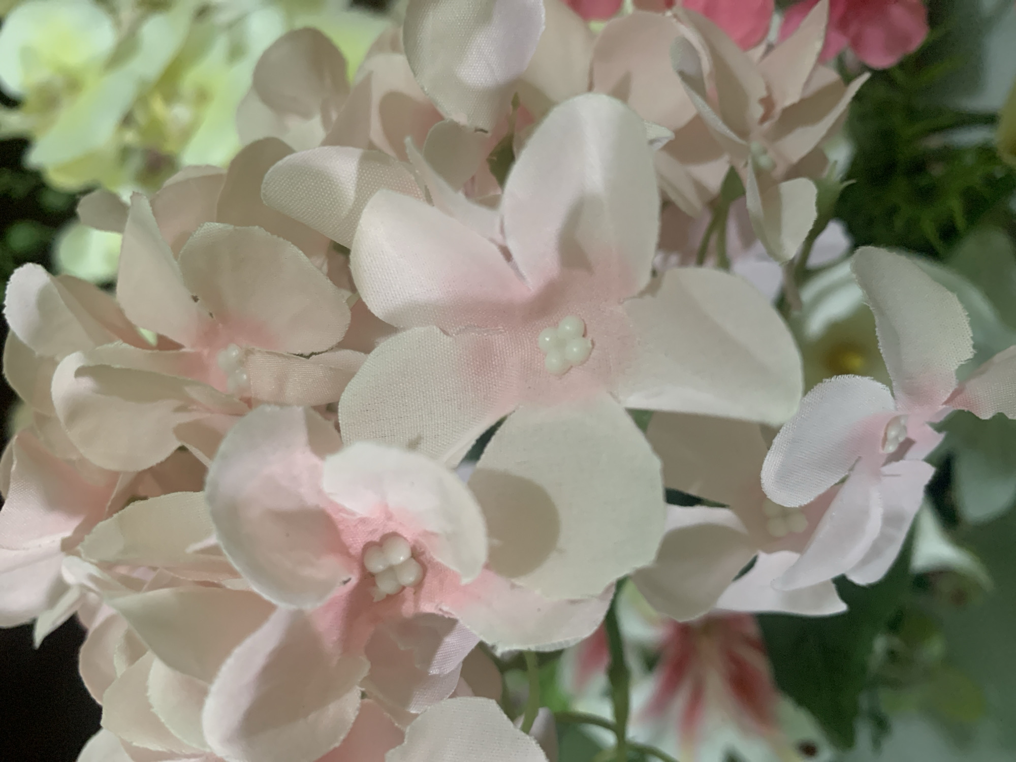 This is a close-up, shallow depth-of-field photograph of artificial pink and white flowers. The flowers appear to be made of fabric with a slight sheen. In the center of some of the flowers, small, pearl-like beads form the stamen. The petals are delicately colored, with a gradient from pale pink at the edges to white closer to the center. The background is out of focus, showing hints of green foliage and possibly other flowers in lighter shades of yellow and pink. The image has a soft, dreamy quality due to the blur. There is no indication of any specific location within Kuala Pembuang, Indonesia; the scene is a generic floral arrangement.