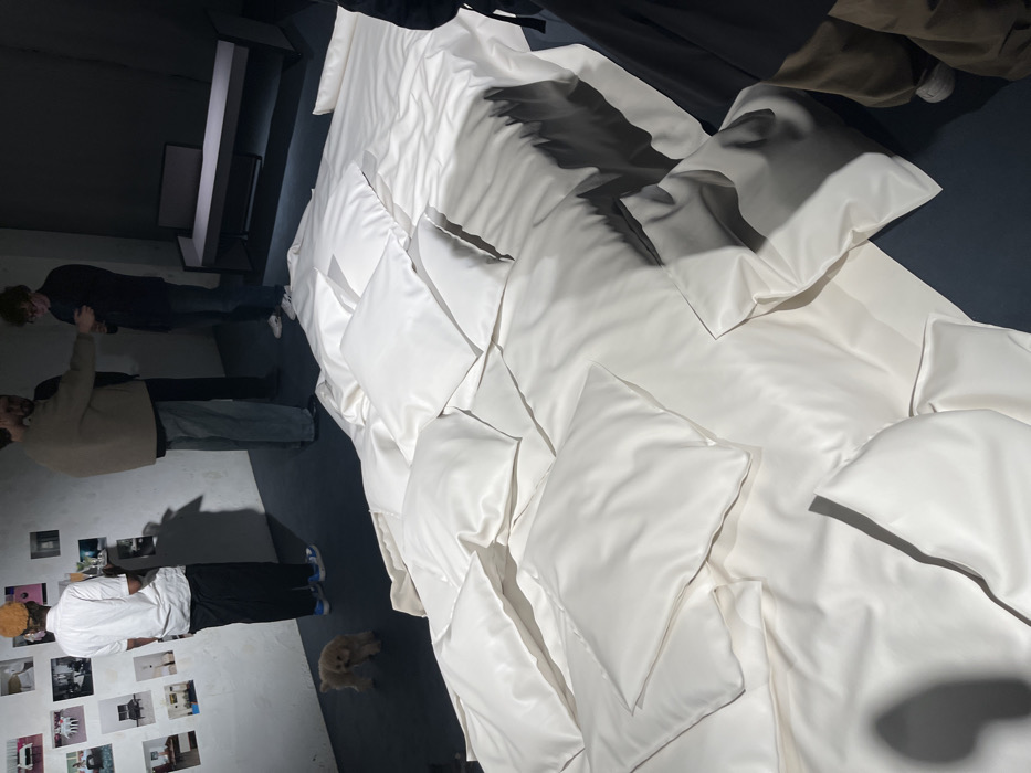The image captures an indoor exhibition space in Paris, France. The foreground is dominated by a sprawling, low art installation resembling a bed, composed of numerous white, possibly vinyl or leather, cushions and mattress-like forms, resting directly on a dark floor.
In the mid-ground on the left, a person with dark skin and light-colored hair, wearing a white short-sleeved collared shirt and black pants, stands facing right. They hold a rectangular tray with two tall glasses containing a yellowish liquid. To their left, a light-colored wall is adorned with a grid of small, unframed photographs depicting interior scenes. A small, light brown, fluffy dog walks on the dark floor between this individual and the wall.
Further right in the mid-ground, two men are engaged in conversation. One man, dressed in a light brown sweater and light-colored pants, stands facing a second man in a dark shirt and dark pants, who appears to be looking down. In the background to the right, a dark, minimalist bench or low console table is visible. The lower section of another person, wearing brown pants and dark footwear, is partially visible on the far right, near the white installation. Directed lighting creates distinct shadows across the floor and the white objects.