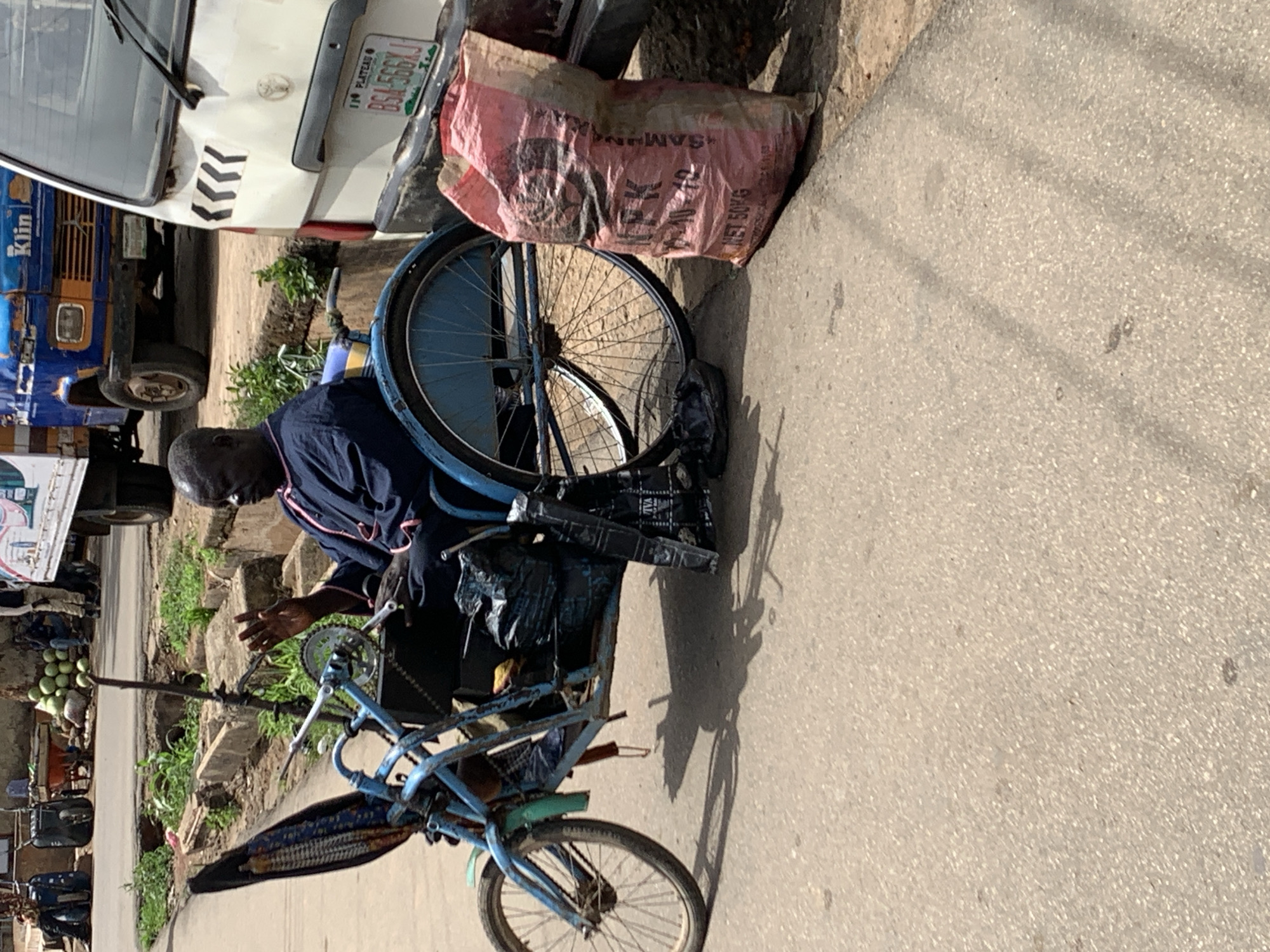 The image shows a man in Jos, Nigeria, repairing a blue bicycle on a paved sidewalk. He is wearing a dark blue long-sleeved shirt. Next to the bicycle is a large, pink sack resting against the back of a white van with a license plate that reads "Plateau BSA-566XJ." In the background, there is a blue and orange truck. The area seems to be a street, with some vegetation in the background.