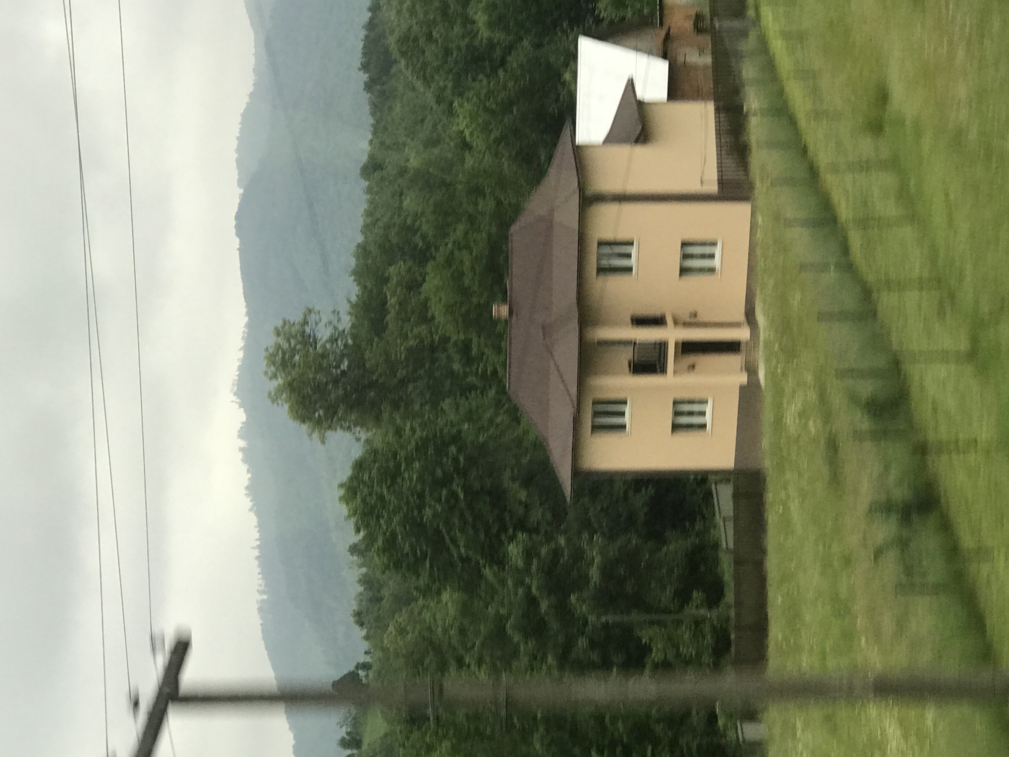 This image captures a rural scene in Krasnoilsk, Ukraine, under an overcast sky. In the foreground, a verdant green grassy field slopes upwards, partially divided by a wire fence running diagonally from the mid-left to the lower right. A blurred utility pole is prominently positioned on the far left. The midground features a two-story, light beige-colored house with a dark brown, multi-pitched roof. The house has multiple rectangular windows with dark frames and a small second-floor balcony with a dark railing centered above a dark main entrance. To the right of the primary residence, a smaller auxiliary building with a distinct white roof and darker-toned walls is partially visible. A dark fence encloses the property surrounding the houses. Immediately behind the buildings, a dense forest of various green deciduous trees stretches across the landscape. The background reveals multiple layers of tree-covered mountains, appearing muted bluish-grey due to atmospheric perspective. The sky is uniformly light grey and cloudy, with faint horizontal lines, likely power lines, traversing the upper portion near the utility pole. No people are present, and no specific actions are depicted.