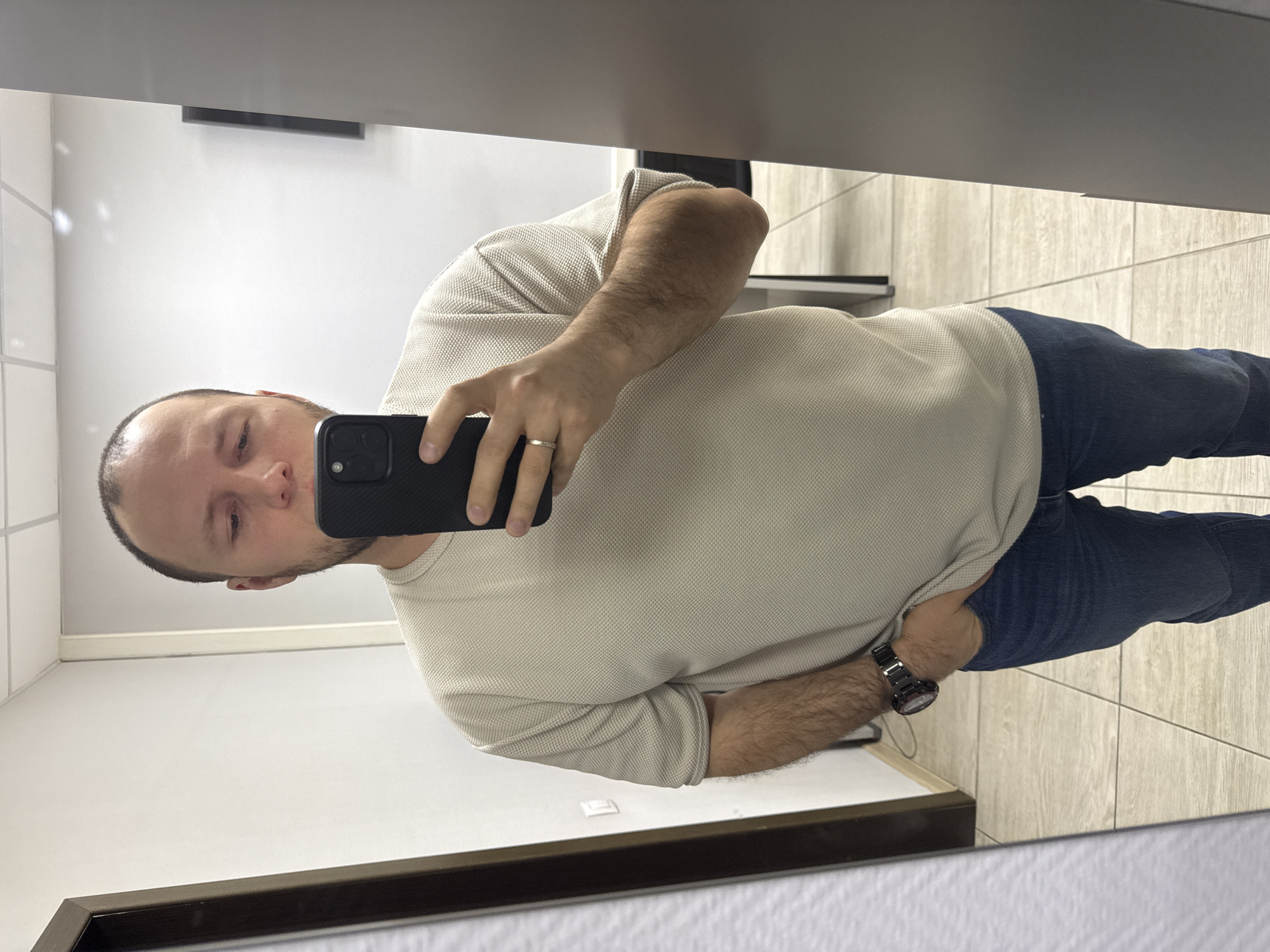 A man is taking a selfie in a mirror. He has a shaved head and is wearing a light-colored long-sleeved shirt and jeans. He is holding a black smartphone in his right hand, and a wedding ring is visible on his finger. He is wearing a dark-colored watch on his left wrist. The background includes a white wall, a dark-framed mirror, and tiled flooring. Based on the interior design and available information, the location could be a restroom or similar facility, but without further details, it is impossible to confirm the exact location in Krasnoyarsk, Russia.
