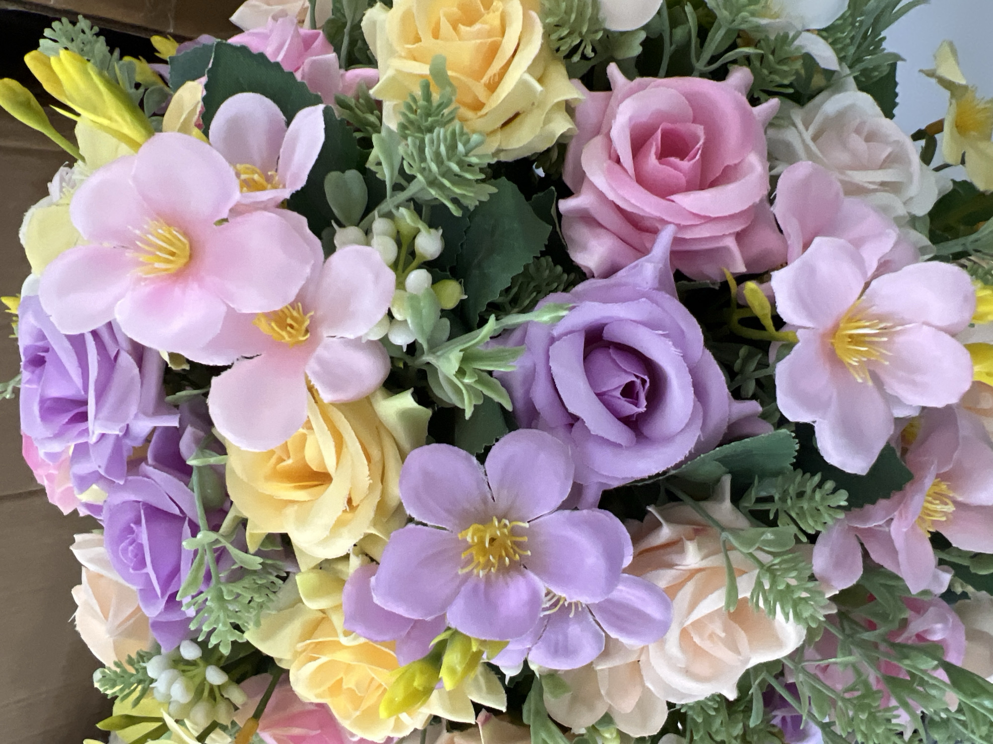 This is a close-up, eye-level, indoor shot of a vibrant bouquet of artificial flowers. The bouquet features a variety of pastel-colored roses in shades of pink, lavender, and cream, interspersed with light purple star-shaped flowers and yellow buds. Green foliage and small white accents add texture and depth to the arrangement. The lighting is soft and even, highlighting the delicate textures and colors of the flowers. There is no discernible background, suggesting the focus is solely on the floral display. The location is specified as Eshanguzar, Uzbekistan.