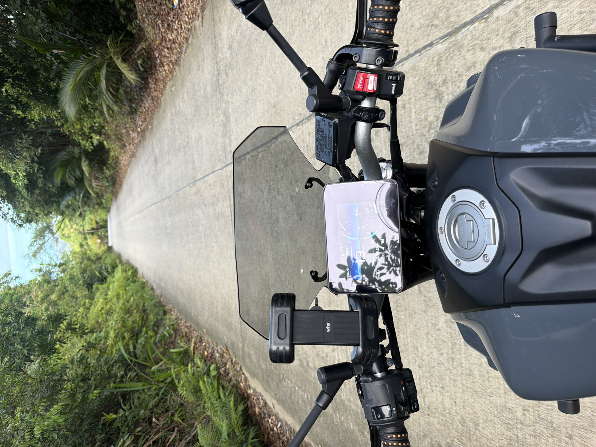 The image is a first-person perspective from a motorcycle traveling on a paved road through a lush, green, tropical environment. The road is empty and appears to be a two-lane street. On either side of the road are dense trees, foliage, and palm fronds, indicative of a tropical setting. The sky is visible at the top of the frame, appearing bright and possibly sunny.
In the foreground, the motorcycle's handlebars and instrument panel are visible. A smartphone is mounted on the handlebars, displaying a digital screen with what appears to be navigation or vehicle data. The motorcycle's windshield is in place, and the front of the fuel tank is also visible. The controls on the handlebars, including buttons and levers, are clearly depicted. The motorcycle's color appears to be a dark gray.
The scene is consistent with a road in a rural or semi-rural area of Baan Tai, Thailand, given the dense tropical vegetation and the style of the road and motorcycle. There are no people visible in the image, and the action is solely the motorcycle's movement forward. The overall atmosphere is one of serene travel through nature.