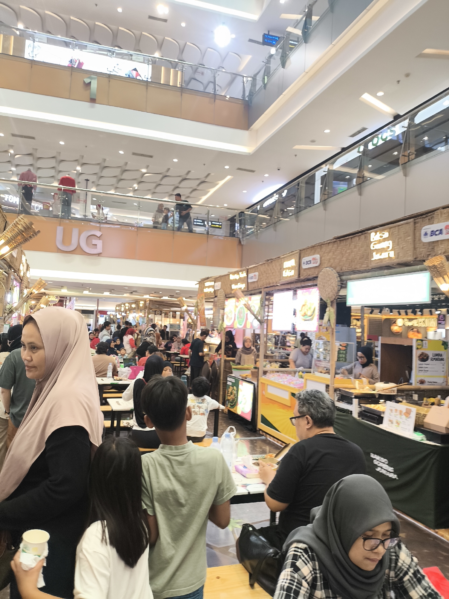 The image depicts a busy interior scene of a shopping mall in Grogol, Indonesia. The scene is multi-level, with escalators and walkways visible on higher floors, showcasing a bright, modern architectural design with ample lighting. The foreground focuses on a food court area where numerous individuals are seated at tables, engaged in dining and social interaction. Several food stalls with prominent signage and display cases are operational. One stall features bamboo-like accents, contributing to an aesthetic that may suggest a market or traditional theme.
In the foreground, to the left, a woman wearing a light pink hijab and dark clothing is prominently featured, holding a white paper cup. To her right, a young boy in a light green t-shirt is facing away from the camera. Behind them, other individuals are also visible, some seated at tables. In the mid-ground, people are queuing at food stalls and walking through the area. The overall atmosphere appears lively and crowded, indicative of a popular commercial hub. The signage in the background includes text in what appears to be Indonesian, such as "UG," "BCA," and food-related names like "Biasa Gising Juara."