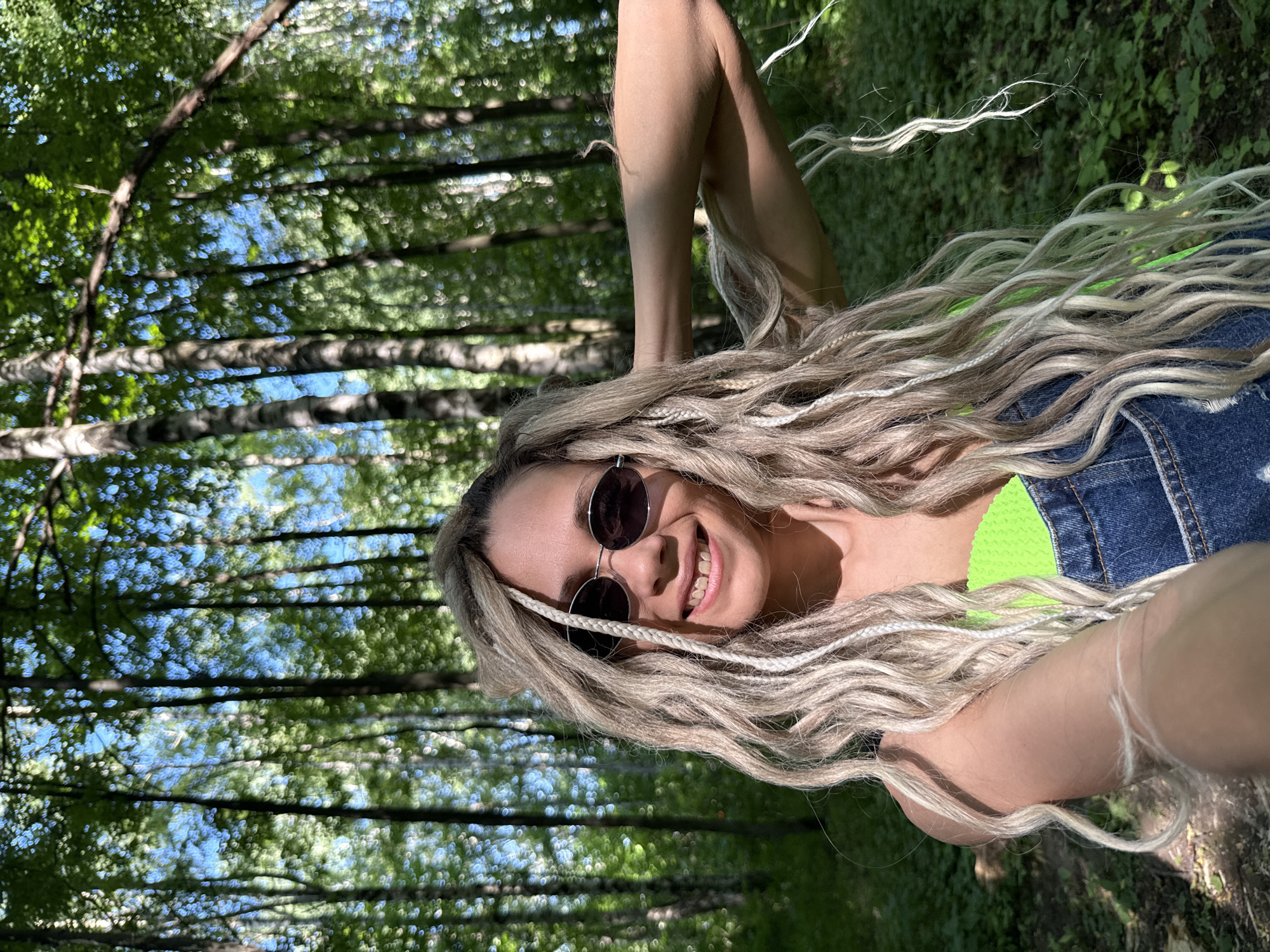 A woman with blonde dreadlocks and sunglasses is taking a selfie in a forest. She is wearing a neon green top and denim overalls. The background shows a dense forest with tall trees and dappled sunlight.