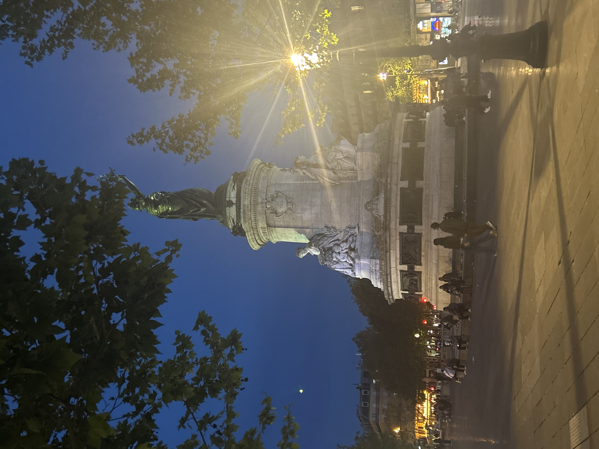 The image shows the Place de la Bastille in Paris, France at night. A large monument stands prominently in the center, likely the Colonne de Juillet. It features a statue at the top and sculptural elements along the base. People are seated and standing around the base of the monument. The night sky is a deep blue. Trees surround the monument, with leaves visible against the sky. A bright street lamp illuminates the monument and surrounding area. Buildings are visible in the background. The perspective is from ground level, looking up at the monument, which is rotated clockwise by 90 degrees. The surrounding pavement appears wet.
