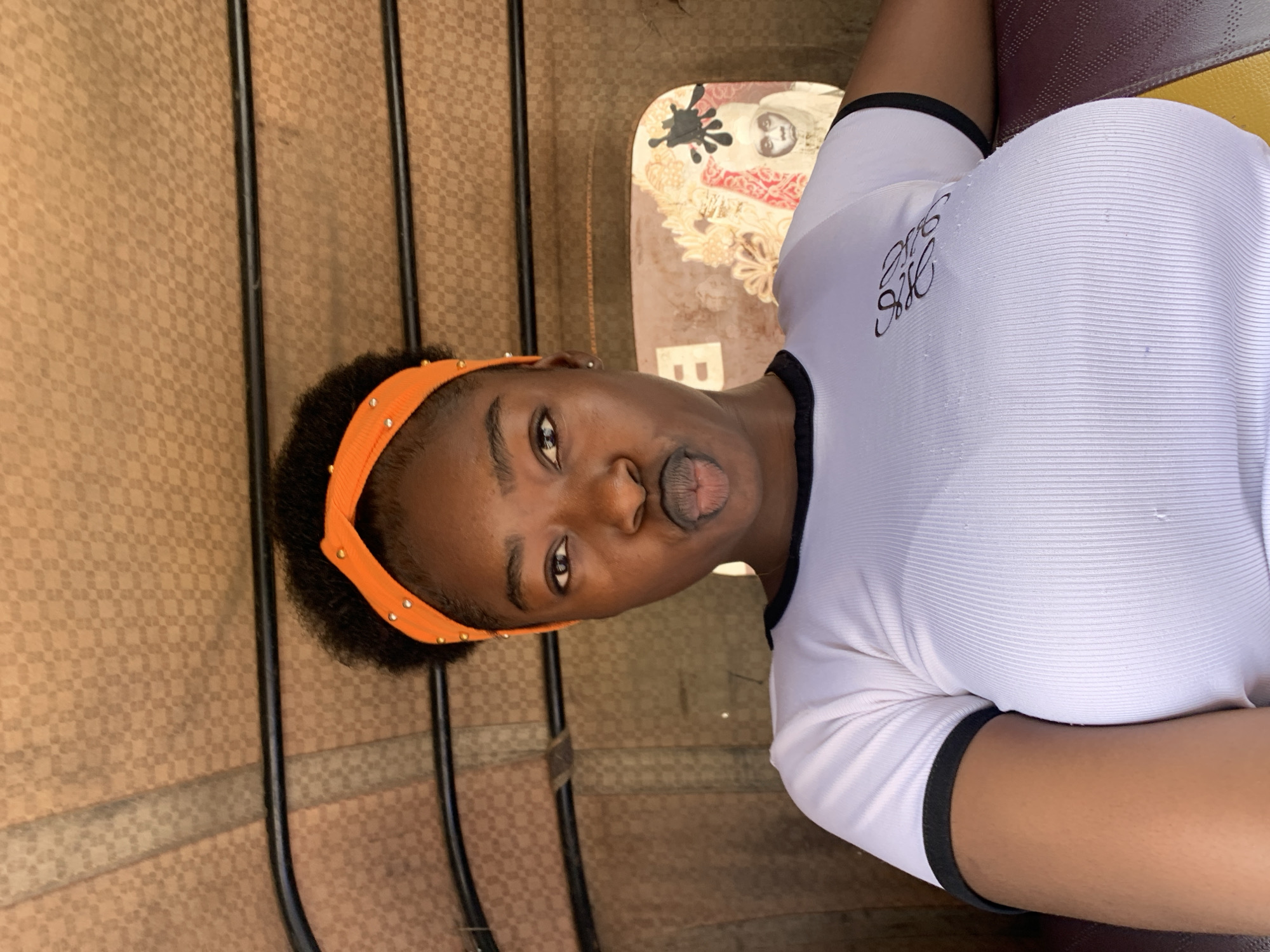 The image depicts a young Black woman inside a vehicle in New Stadium, Nigeria. The woman is wearing a white, short-sleeved shirt with black trim and an orange headband with small, round embellishments. She is making a puckered face. The background includes a patterned wall, metal bars, and a painted graphic on the vehicle's interior. A brown-colored textured surface visible near the bottom of the image suggests a seat.
