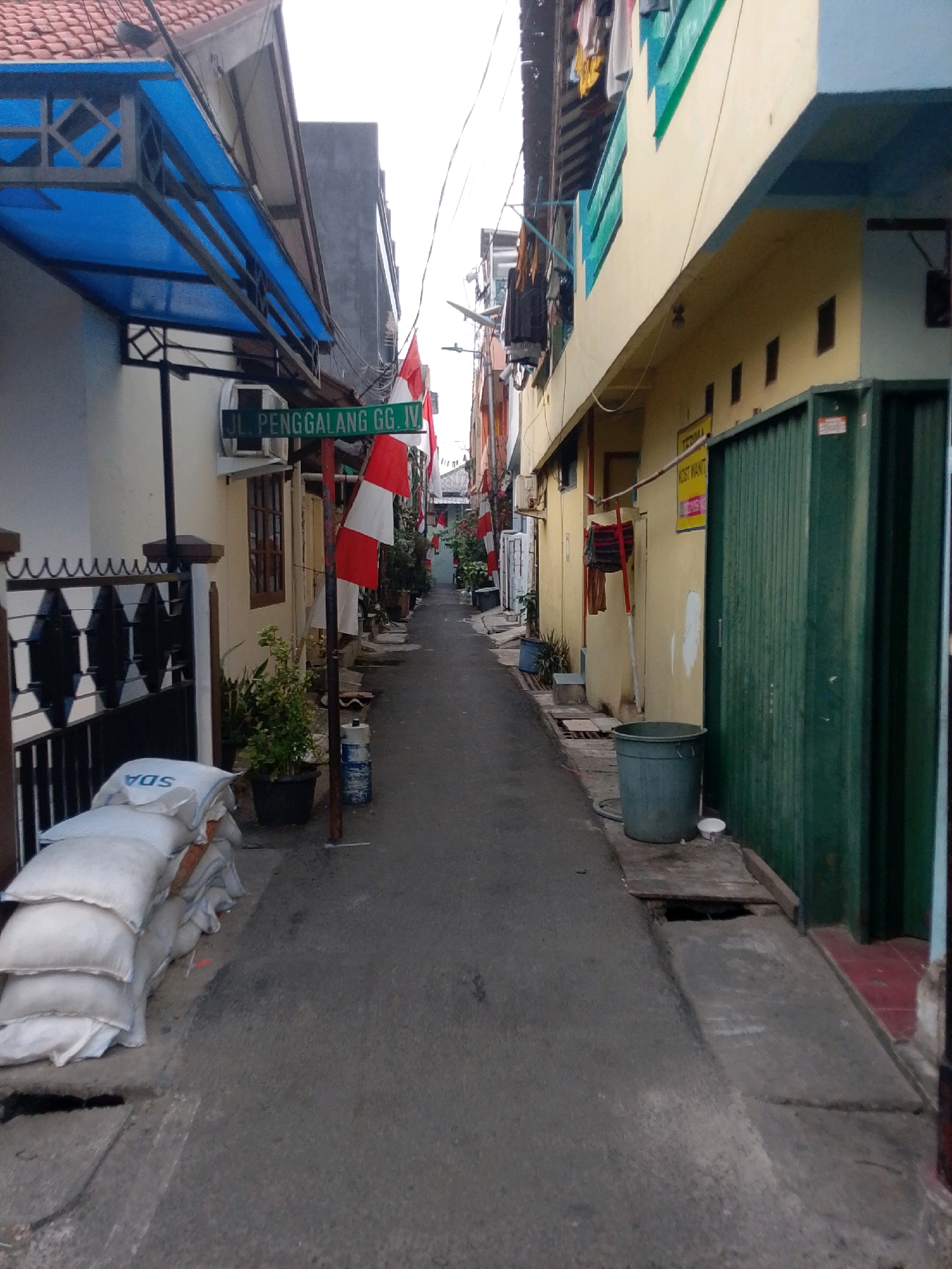 The image captures a narrow alleyway in East Jakarta, Indonesia, marked by the street sign "JL. PENGGALANG GG. IV". The alley is lined with buildings on both sides, some appearing residential and others commercial. On the left, a white building features a blue awning and a black metal fence. Stacked white sacks, possibly containing construction materials, are positioned near the fence. Further down the alley on the left, more buildings are visible, adorned with Indonesian flags.
On the right side of the alley, a yellow building has a green corrugated metal gate and a sign that reads "TERIMA KOST WANITA" (Women's Boarding House). A large green plastic trash bin stands near the gate, and a small white cup is placed on the ground beside it. Clothes hang on a line above the yellow building, and laundry is also visible on balconies. The alleyway itself is paved with asphalt. The scene appears to be a typical street view in a densely populated urban area.