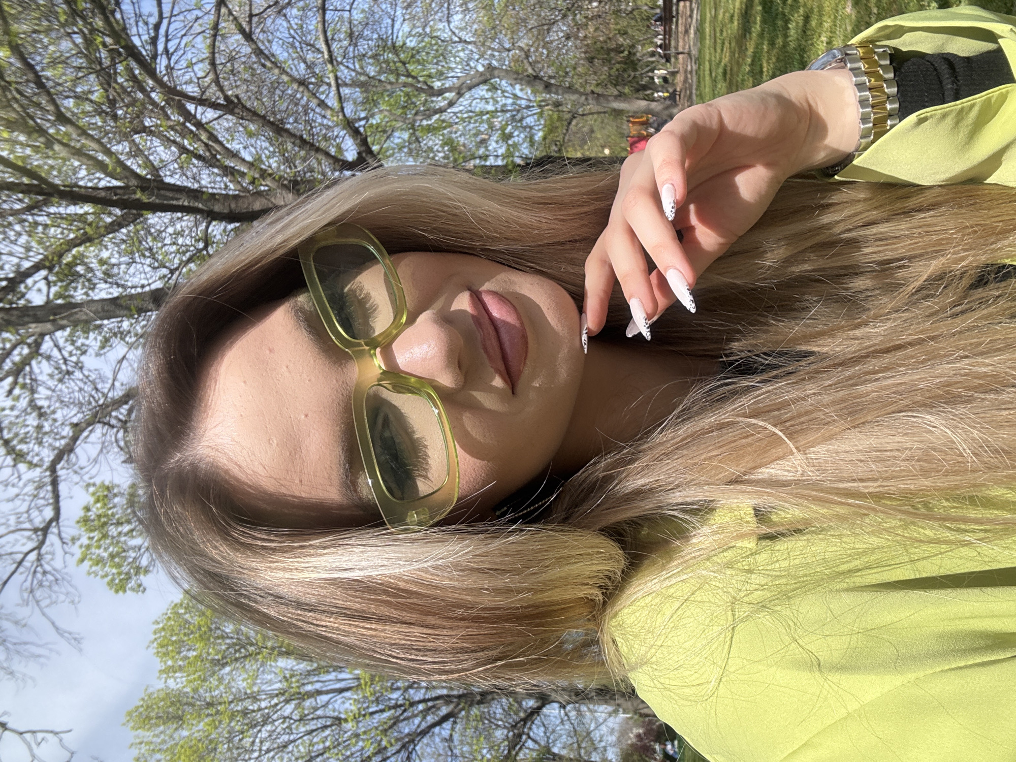A close-up outdoor selfie, taken in Plovdiv, Bulgaria, features a person, likely female, from the chest up, posing on a bright, sunny day. The subject has long, flowing blonde hair that frames her face and extends over her shoulders. She is wearing distinct light yellow-green rectangular sunglasses, which reflect the surrounding tree branches and sky. Her lips are slightly parted and appear glossy.
She is dressed in a vibrant lime green or neon yellow long-sleeved top or jacket. On her left wrist, a two-tone watch with both silver and gold links is visible. Her left hand is raised near her face, showcasing her long, almond-shaped nails, which are painted white with a delicate black speckled or dotted design.
The background reveals a clear, light blue sky above a network of tree branches. Many of the branches are budding with fresh, light green leaves, indicating that the photo was taken in early spring. The setting appears to be a park or an outdoor green space, with hints of distant greenery in the background. The bright sunlight suggests it's likely midday or early afternoon. The overall impression is one of casual outdoor enjoyment. There is no visible text in the image.