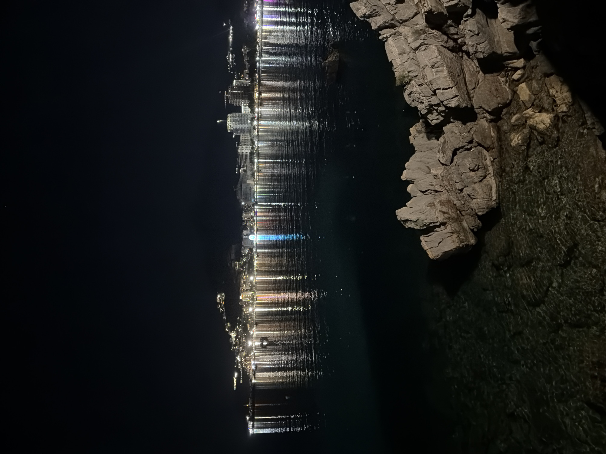 The image depicts a nighttime view of the Budva Riviera in Montenegro, with the city's skyline illuminated against the dark sky. In the foreground, rough, light-colored rocks are visible at the edge of the dark water. The water itself is calm, reflecting the colorful lights of the buildings and a Ferris wheel on the city's waterfront. The buildings appear to be a mix of hotels and residential structures, situated on a hillside that slopes down towards the sea. The lights are a mix of warm white, yellow, and some blue hues, creating a vibrant and festive atmosphere. A small, dark boat is present on the water in the middle ground. The sky is a deep, starless black, emphasizing the glow of the city.