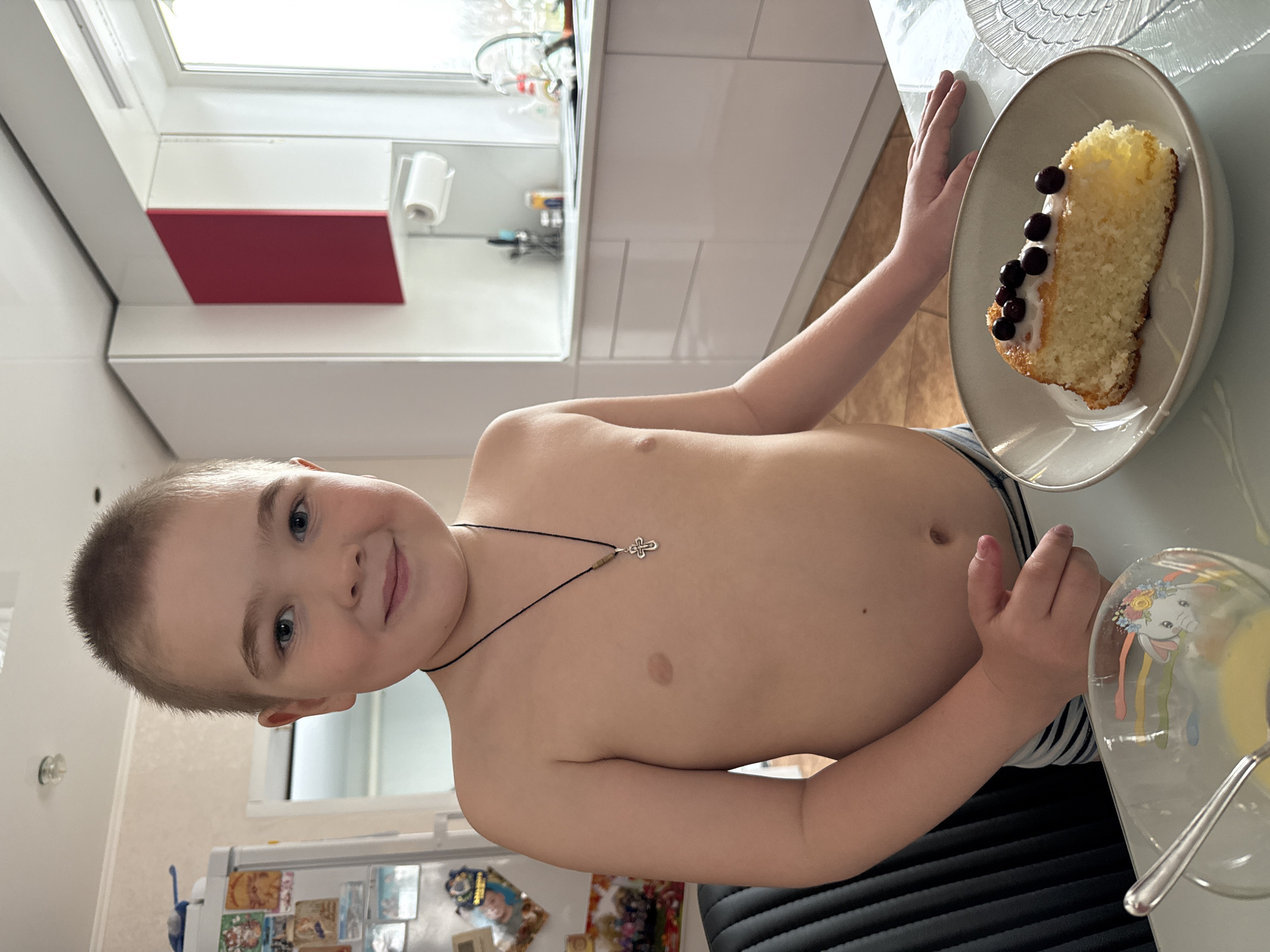 A young, fair-skinned boy with short blonde hair and blue eyes stands shirtless in what appears to be a kitchen. He is smiling towards the camera. He wears a necklace with a small silver cross pendant. To his right, there is a slice of cake topped with cherries and a glass with a yellow liquid inside, sitting on a white countertop. The background reveals a modern kitchen setting with white cabinets, a window, and a red panel on one of the cabinets. A refrigerator with magnets and photos is visible. The location in the city of Zhigalki, Russia, is not verifiable from the visual information provided.