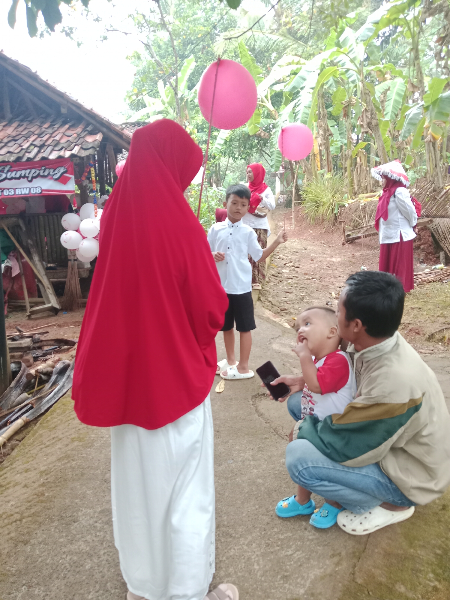 In Bojongmengger, Indonesia, a group of people are gathered outdoors, possibly for a celebration. A woman in a red hijab and white skirt stands facing away from the camera, obscuring her face. To her right, a young boy in a white shirt and black shorts looks towards the viewer. Behind them, a woman in a red headscarf and white top, and another woman in a red headscarf and a traditional conical hat, are visible. In the foreground, a man is crouching, holding a young child who is wearing a red and white shirt. The man is holding a black mobile phone. A few pink balloons are floating in the background. The environment appears to be a rural or semi-rural setting, with vegetation including banana trees visible. A wooden structure with a tiled roof, possibly a building or stall, is present on the left side of the image.