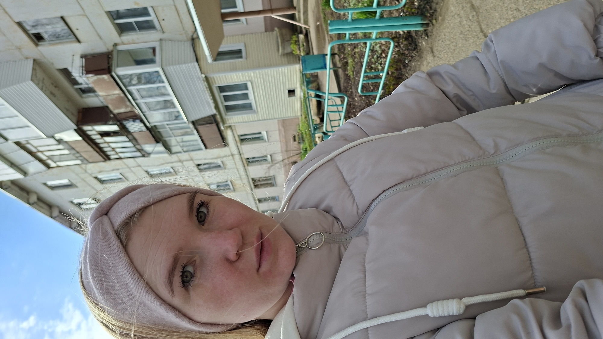 A young woman with a pink headband and a light gray puffy jacket is facing the camera, the image is rotated counterclockwise. She has fair skin, blonde hair, and green eyes. The zipper of her jacket is partially visible and the drawstring hangs loose. Behind her, the corner of a building with multiple windows and balconies under a blue sky is visible. A playground with green metal structures is also visible in the background. The location is Chaikovsky, Russia.