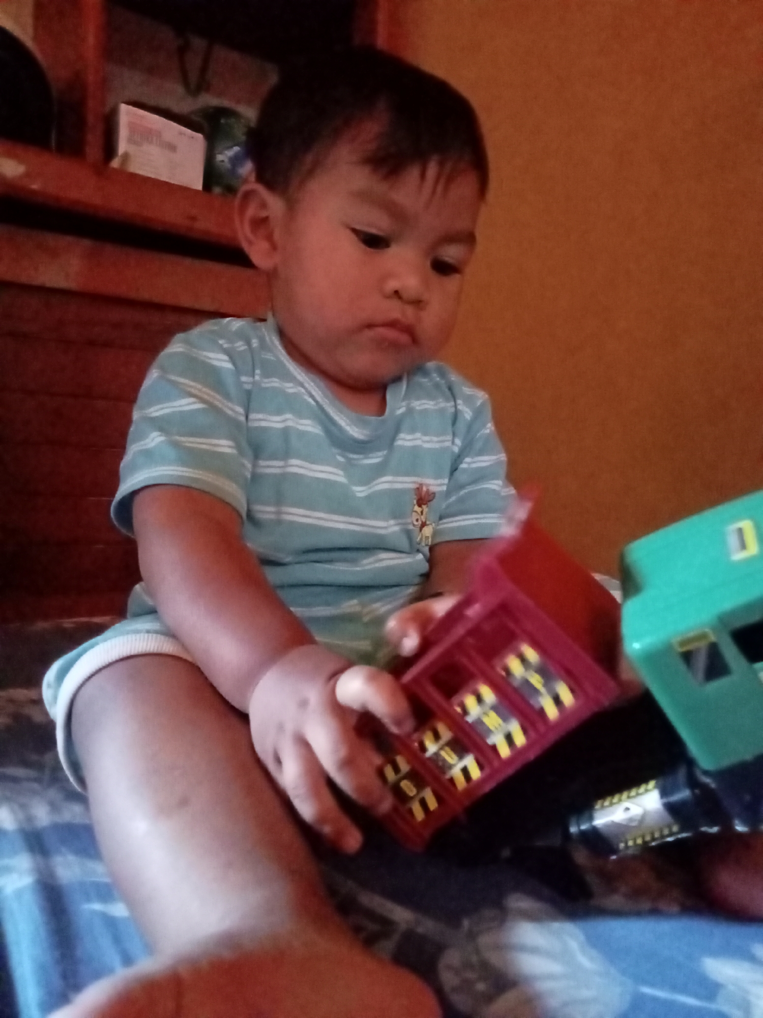 A young child is sitting on a bed, playing with toy trains. The child is wearing a light blue and white striped t-shirt and white shorts. The child's left hand is holding a red toy train car with yellow windows, and the right hand is reaching for a green toy train car. The background shows a wooden shelf and a wall. The setting is likely a bedroom. The image appears to be taken in Pancurwening, Indonesia.