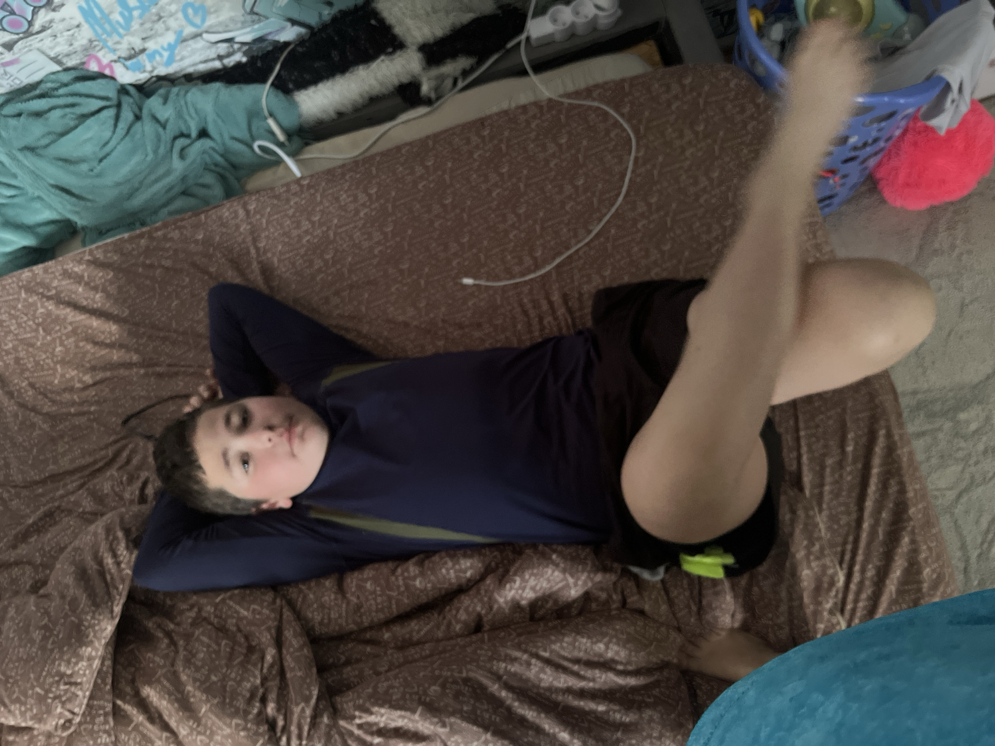An overhead shot captures a young person, likely a boy, lying on their back on a bed covered with brown patterned bedding. He has short dark hair and a neutral facial expression, with his head resting on his clasped hands. He is wearing a dark blue long-sleeved shirt with a light-colored (possibly olive green) stripe on the sleeves and dark shorts. One leg is bent at the knee, while the other is extended upwards towards the top right of the frame, suggesting a relaxed or playful pose.
The setting appears to be an indoor room, possibly a bedroom, characterized by a mix of functional and personal items. In the upper left corner, a teal crumpled blanket or piece of fabric displays faint, illegible scribbled text or doodles. Beneath it, another light-colored object (perhaps a poster or paper) also shows faint, unreadable white text/doodles. Next to these, a black and white patterned rug or blanket is visible. A white cable or cord snakes across the bed and onto the floor from the upper left. In the upper right, a blue plastic laundry basket contains various items, including a grey garment, and a bright pink fuzzy object rests beside it. A white power strip with multiple outlets is visible on a surface behind the blue basket. A light blue blanket or fabric partially covers the bottom right corner of the frame. The overall impression is one of casual relaxation in a lived-in space. The location context is Vesyoloye, Russia, though the image itself offers no specific visual cues to confirm this.