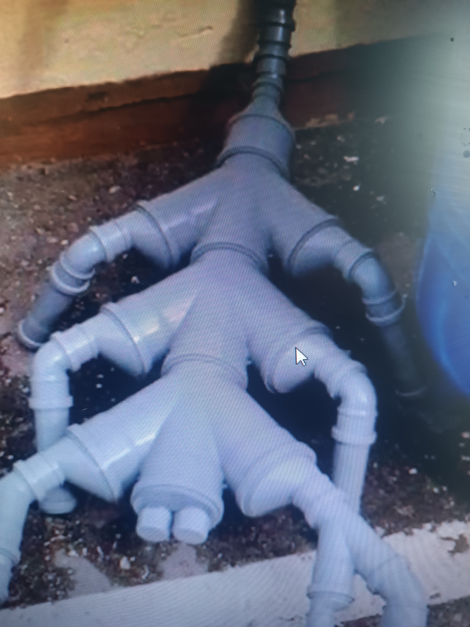 The image displays a complex arrangement of light blue polyvinyl chloride (PVC) pipes, resembling a multi-pronged structure or a system of interconnected branches. The pipes are joined at various angles, creating a visually intricate network. The main pipe enters from the top center, then splits into several branches, which further divide. The pipes are situated on a rough, gravelly surface, with a darker, horizontal element visible in the upper portion of the image, possibly a wall or a foundation. The lighting appears to be somewhat diffused, casting subtle shadows. The overall impression is of an external plumbing or drainage system, possibly in a construction or maintenance area. The location in Klyuchi, Russia, is not visually apparent from the provided image.