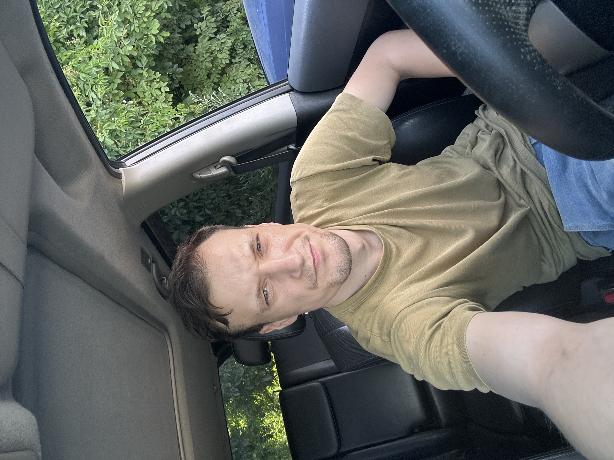 A man is sitting in the driver's seat of a car, taking a selfie. He has short brown hair and a light beard. He is wearing a short-sleeved olive green t-shirt and blue jeans. The car's interior is visible, including black leather seats and a sunroof. Outside the car, green foliage can be seen through the windows. The scene appears to be in a natural setting, possibly a rural area outside of Shatava, Ukraine. The lighting suggests it is daytime.