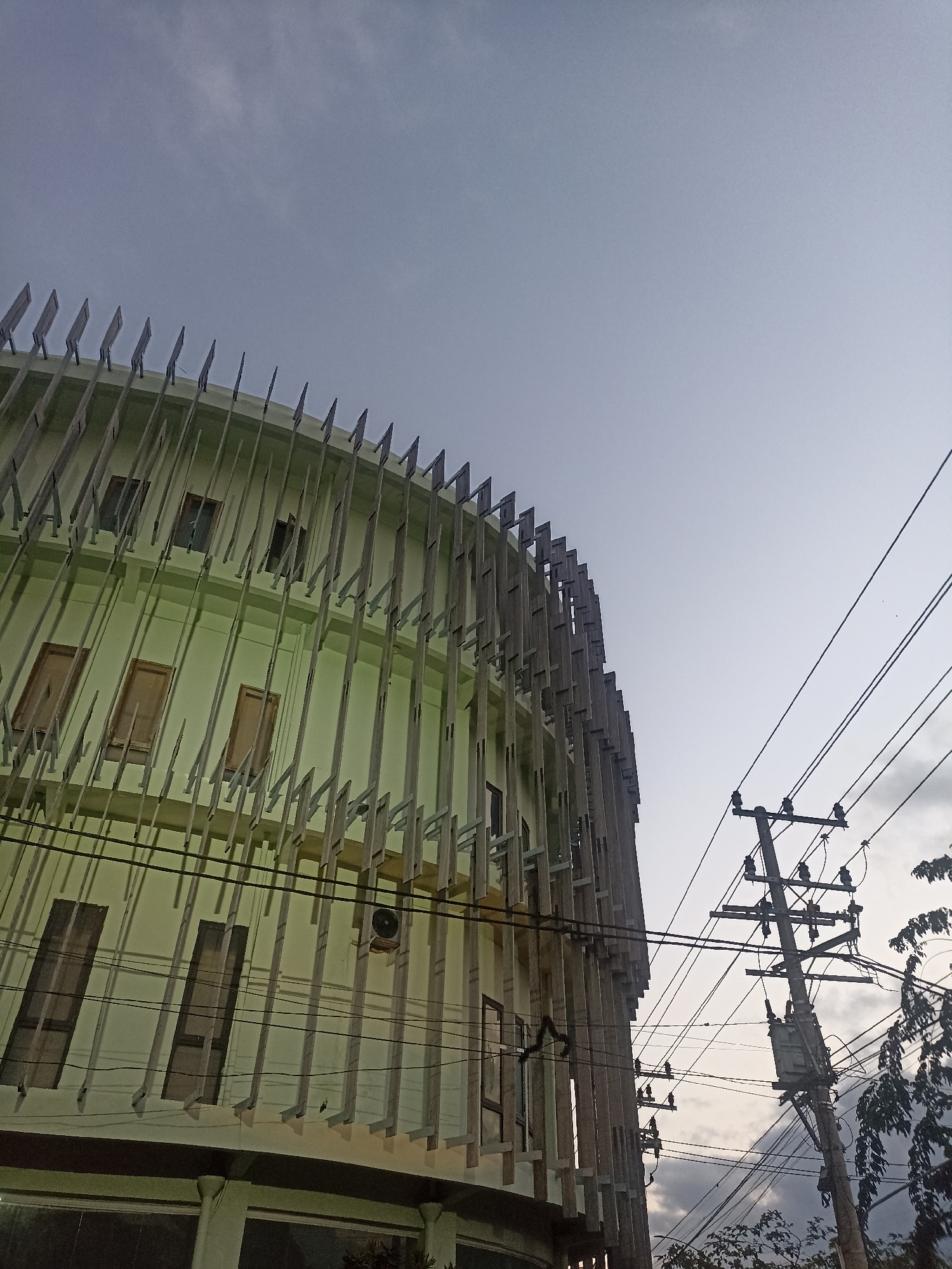 This low-angle, outdoor photograph captures a building with a distinctive, modern facade, located in Mojopanggung, Indonesia. The building is primarily pale green and features a curved exterior adorned with vertical wooden slats arranged in a staggered pattern. Several rectangular windows, some with dark panes, are visible on the building's surface. An air conditioning unit is mounted on the wall. To the right of the building, a wooden utility pole with multiple wires and a transformer box is prominently featured. The sky above is a pale, overcast gray. The image appears to have been taken during daylight hours, possibly in the late afternoon or early morning, given the soft lighting. No people are visible in the frame. The overall impression is of an urban or semi-urban environment.