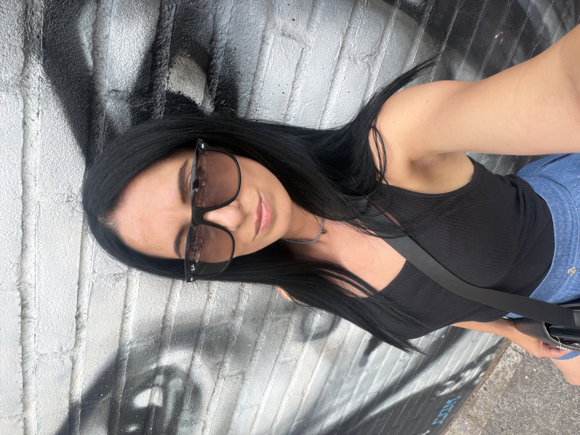 A medium shot selfie captures a woman with long, dark hair, wearing black sunglasses and a black tank top, standing against a graffiti-covered brick wall. She is also wearing a blue polka-dotted skirt and a black purse with a strap across her chest. The woman is looking at the camera and smiling slightly. The background is a brick wall with black and white graffiti art. The image appears to be taken outdoors in an urban setting, possibly in Corsico, Italy, given the context. The lighting is bright, suggesting it is daytime.