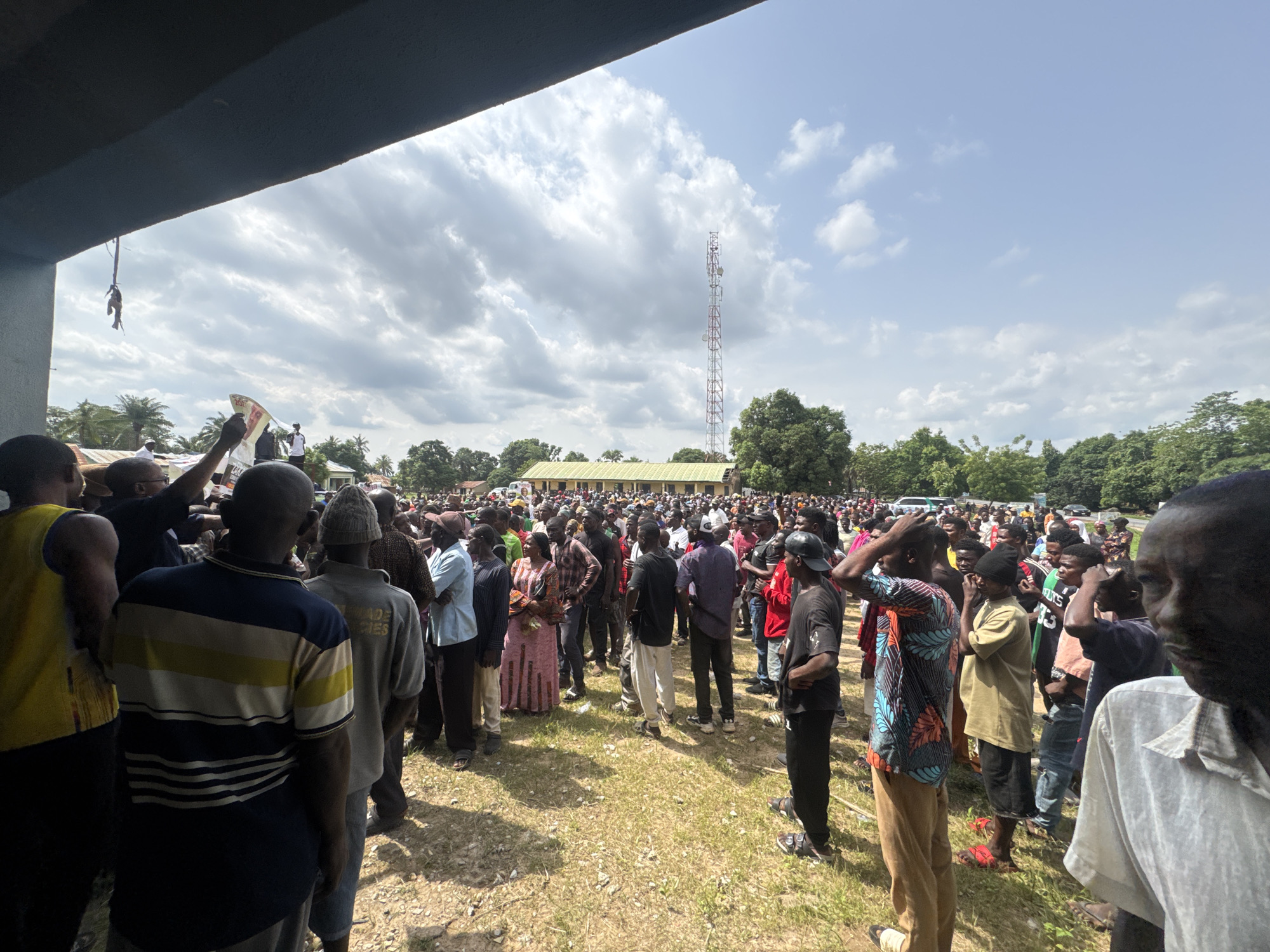 The image depicts a large crowd of people gathered outdoors in Akwanga, Nigeria. The crowd consists of people of varying heights, dressed in diverse clothing. They are standing on a grassy and gravelly surface. The view is partially obstructed by a concrete overhang at the top left.
In the background, there is a building with a light-colored roof and a tall telecommunications tower. The sky is partly cloudy. Some people in the crowd are looking towards a point of interest outside the frame of the image.