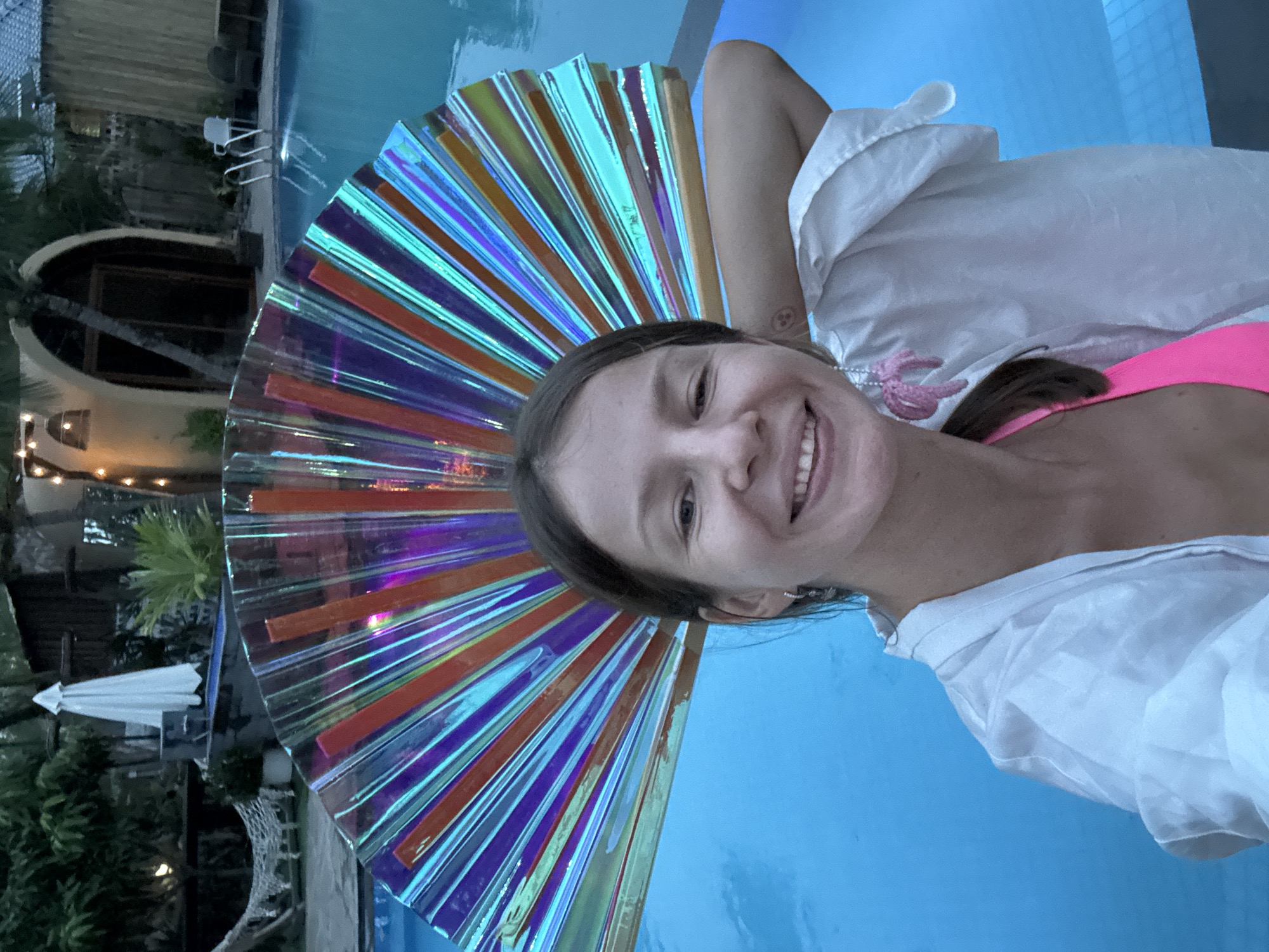 The media file is a portrait-oriented, low-angle selfie of a young woman lying down by a swimming pool. The woman has light brown hair and is smiling at the camera, wearing a pink bikini top and a white sheer shirt. A large, fan-shaped iridescent accessory, with strips of orange, purple, and blue, is positioned behind her head, creating a halo effect.
The setting appears to be a resort or hotel in Mũi Né, Vietnam. In the background, to the left, a circular building with an arched entryway and illuminated windows is visible, surrounded by lush green vegetation and palm trees. A white folded umbrella and patio furniture are also present near the pool. The pool itself is a vibrant blue, reflecting the bright sky, suggesting it is daytime, likely late afternoon or early evening given the lighting. The overall mood is relaxed and happy, with the woman enjoying her vacation. There is no visible text in the image.