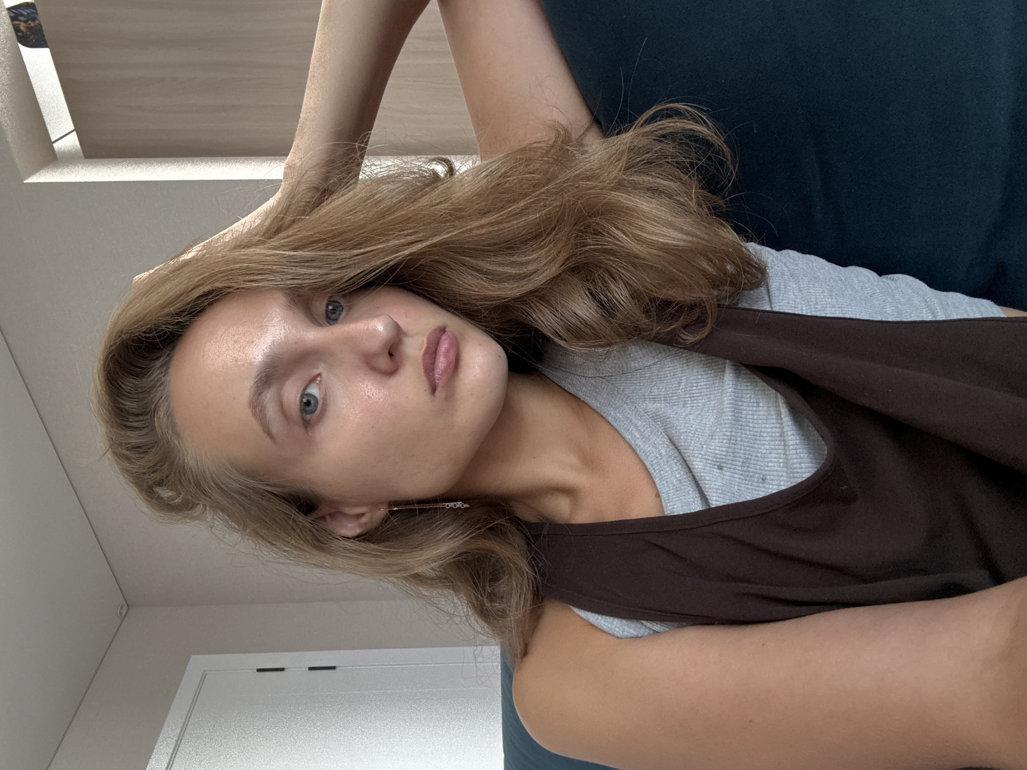 The image shows a young woman with blonde hair and blue eyes, taking a selfie indoors. She is wearing a grey tank top under a brown vest. Her left arm is resting on her head, and her right arm is resting on her side. The background appears to be a living room with a sofa and a door. There is no indication of the city of Подшивалово, Russia.