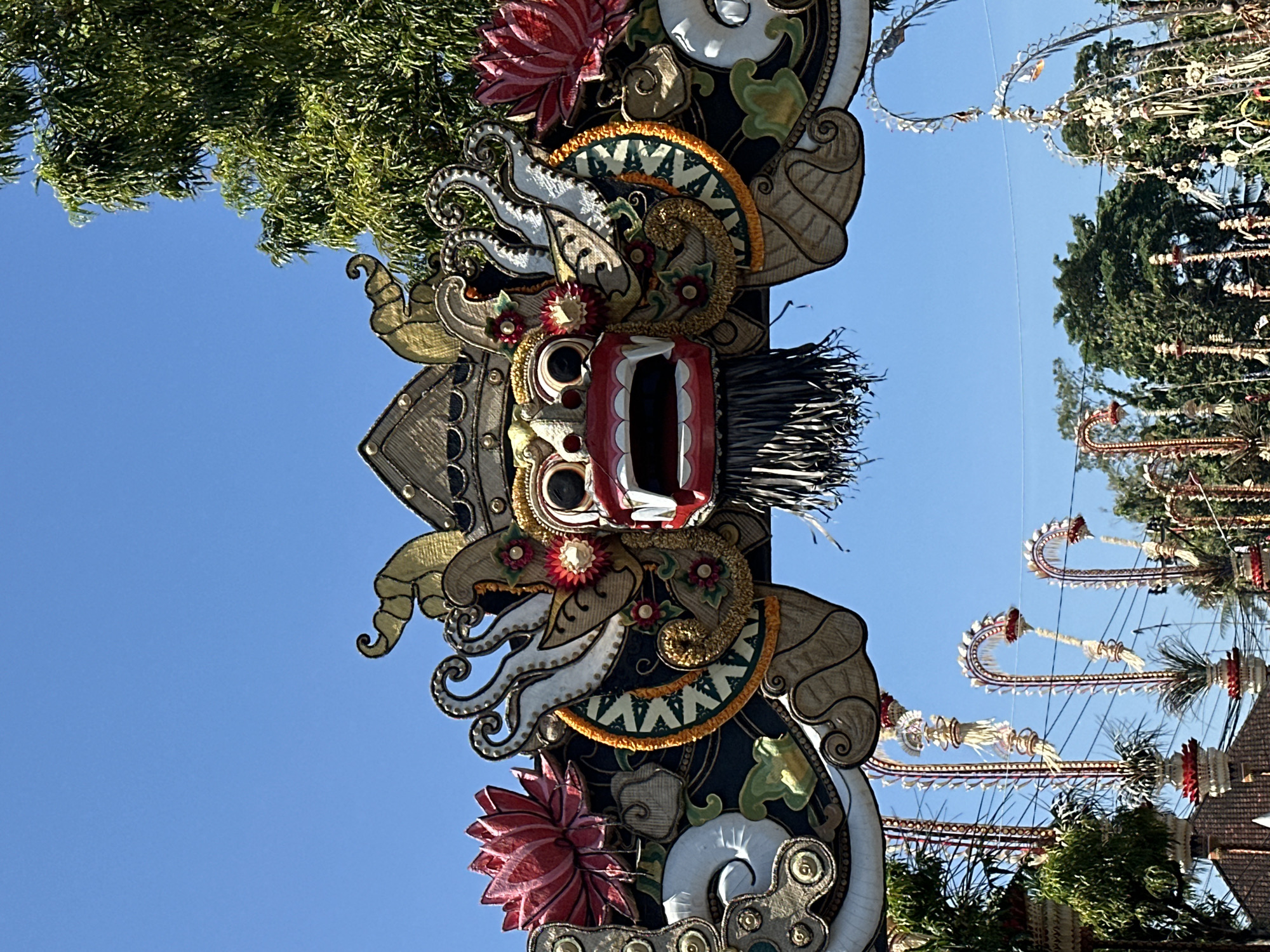 The image shows a close-up of an ornate, traditional Balinese gate in Sukawati, Indonesia, under a clear blue sky. The gate is decorated with a large, fierce-looking carved or sculpted head with wide eyes, an open mouth with sharp teeth, and red lips, resembling a demon or mythological creature. It is adorned with intricate details, including gold accents, red lotus flowers, and what appear to be woven or textured materials. Surrounding the central face are elaborate, swirling decorative elements in white, gold, and dark green, featuring patterns and smaller floral motifs. Below the gate, several tall, decorative poles with arched tops and ornate tassels are visible, suggesting a festive or ceremonial occasion. Green foliage from trees is present in the background and to the right of the gate, and electrical wires are faintly visible among the decorations. The lower portion of the image shows parts of buildings with tiled roofs and more of the decorative poles. The overall impression is one of vibrant cultural artistry and tradition.