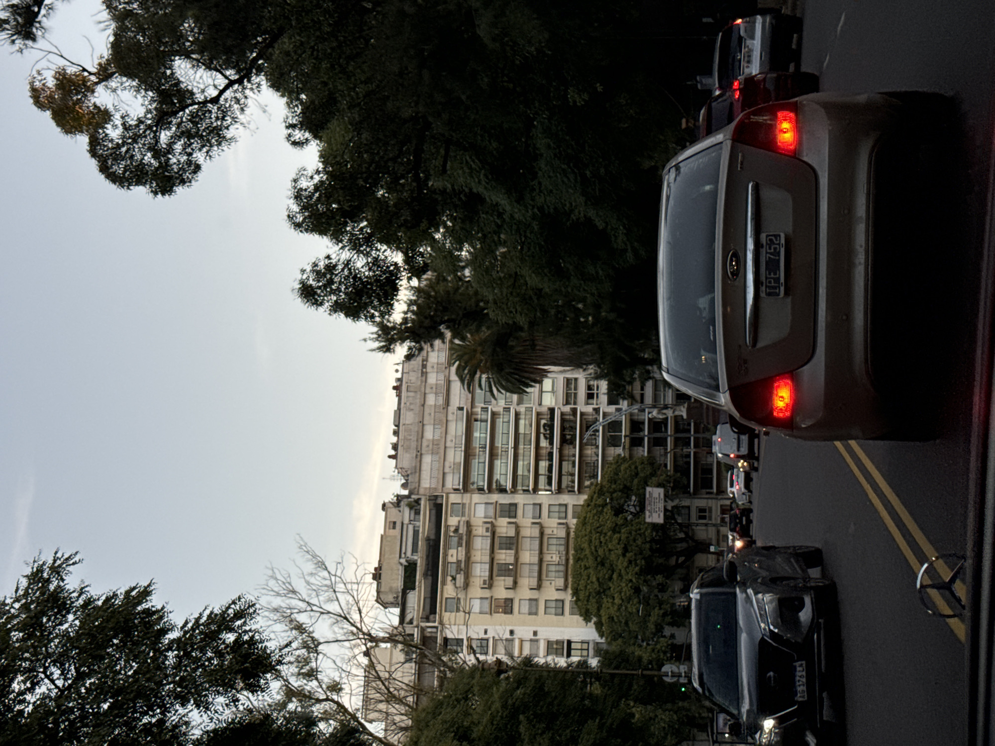 The image depicts a street scene in Buenos Aires, Argentina, showcasing heavy traffic with multiple cars lined up on the road. The license plates visible suggest the vehicles are registered within the country. High-rise apartment buildings line the street, partly obscured by trees. The sky is overcast, indicating a cloudy day. The vehicles are stopped or moving slowly, likely due to congestion.